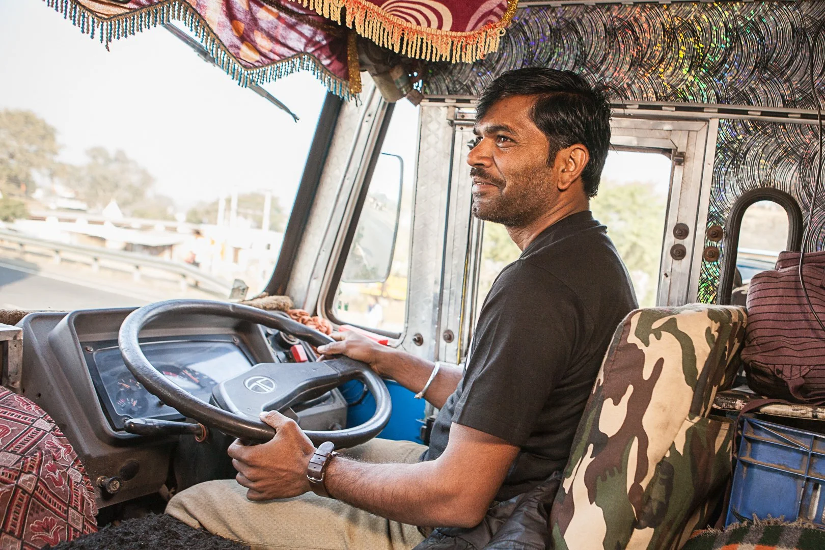 A man driving a truck, seated at the steering wheel with a smile, with a view of the road outside through the front window.