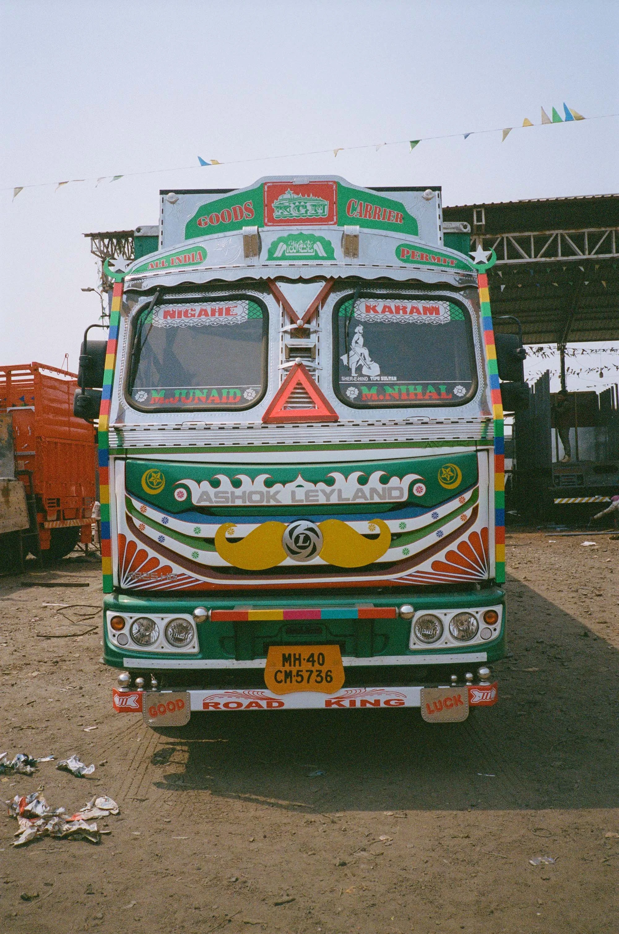 Colorfully decorated truck with various patterns, symbols, and writings, including 'Ashok Leyland' brand and license plate MH-40 CM-5736, parked on a dirt ground.