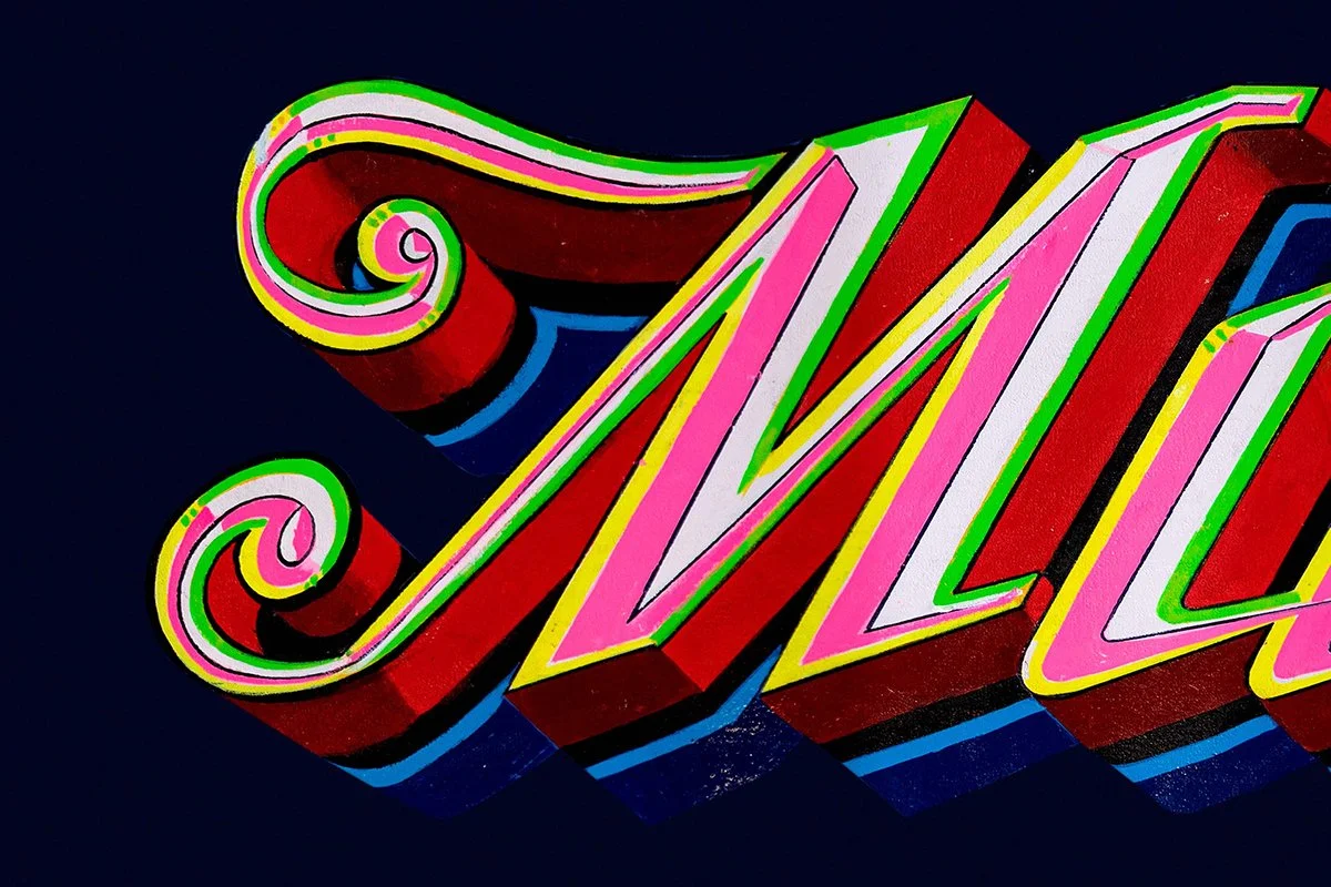 Colorful neon sign spelling out "Ma" with decorative swirls, set against a dark background.