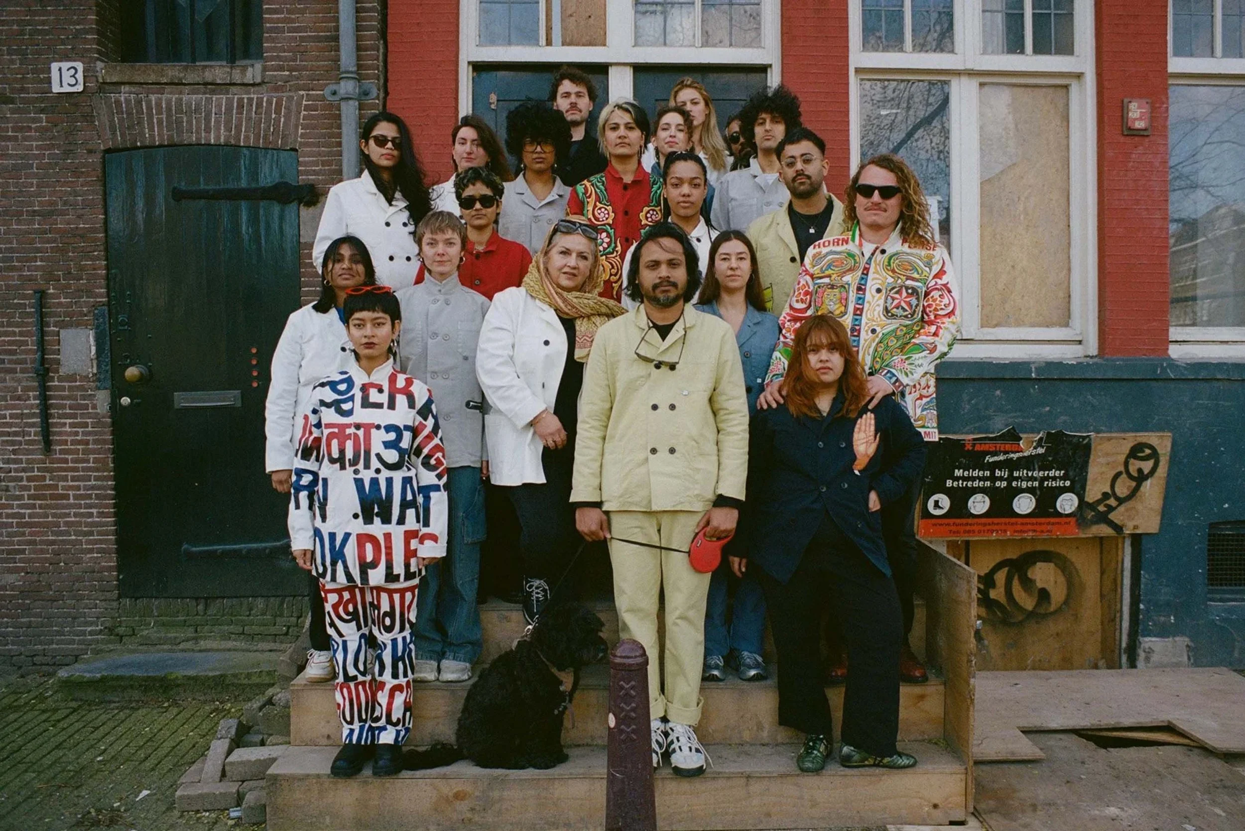 A diverse group of people standing on steps outside a brick building, some wearing colorful and patterned clothing, accompanied by a black dog.