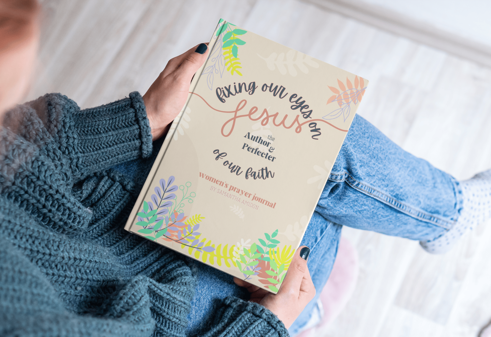 A woman wearing a cozy blue knitted sweater and jeans sitting on a light wooden floor, holding a book titled 'Fixing Our Eyes on Jesus: The Author & Perfector of Our Faith Women's Prayer Journal by Samantha Amidon' with a cover decorated with colorful leaves.