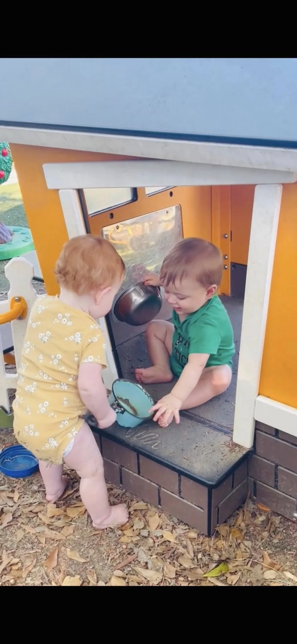 Two young children playing outdoors in a sandbox area of a play structure. One child with red hair is standing, while the other with brown hair is sitting inside the playhouse, both engaging in pretend play with a metal pot and a small blue bowl. There are fallen leaves on the ground.
