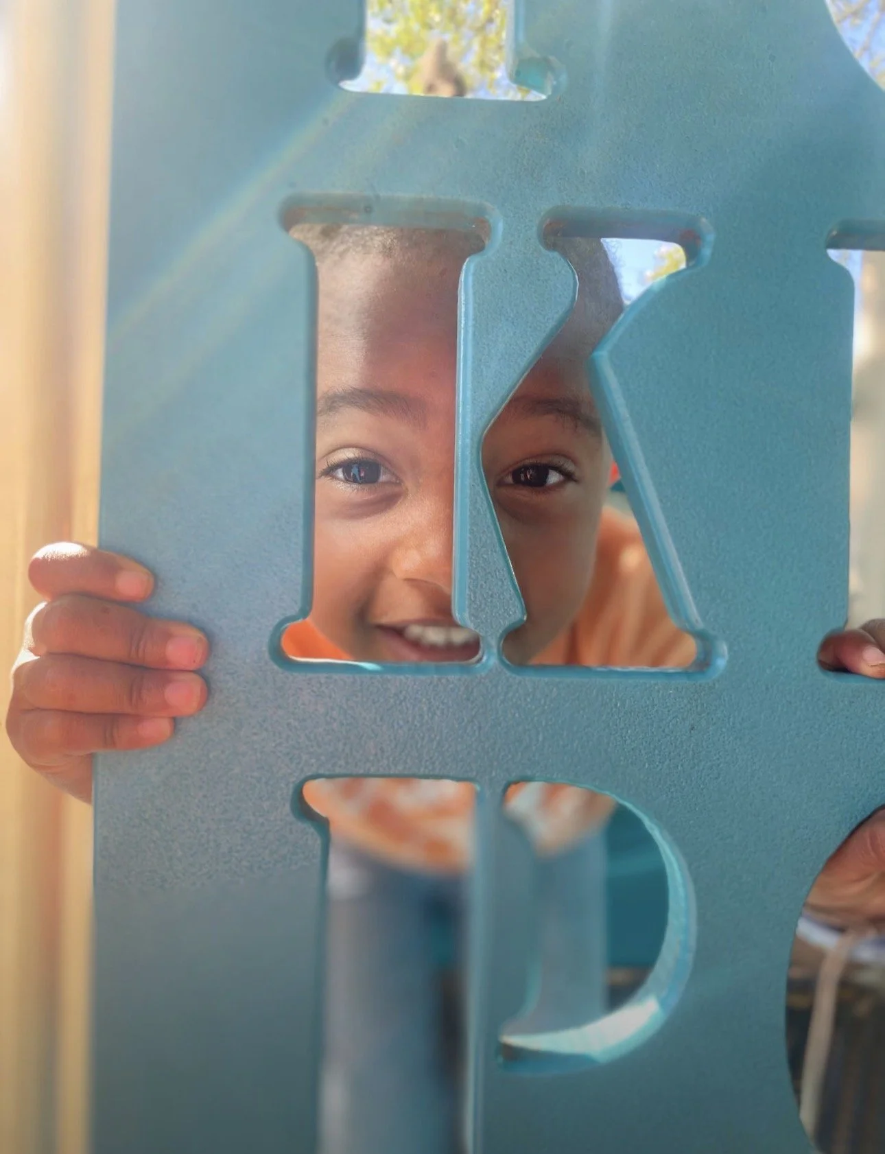A child peering through a blue plastic cutout in the shape of a letter, smiling and looking playful.