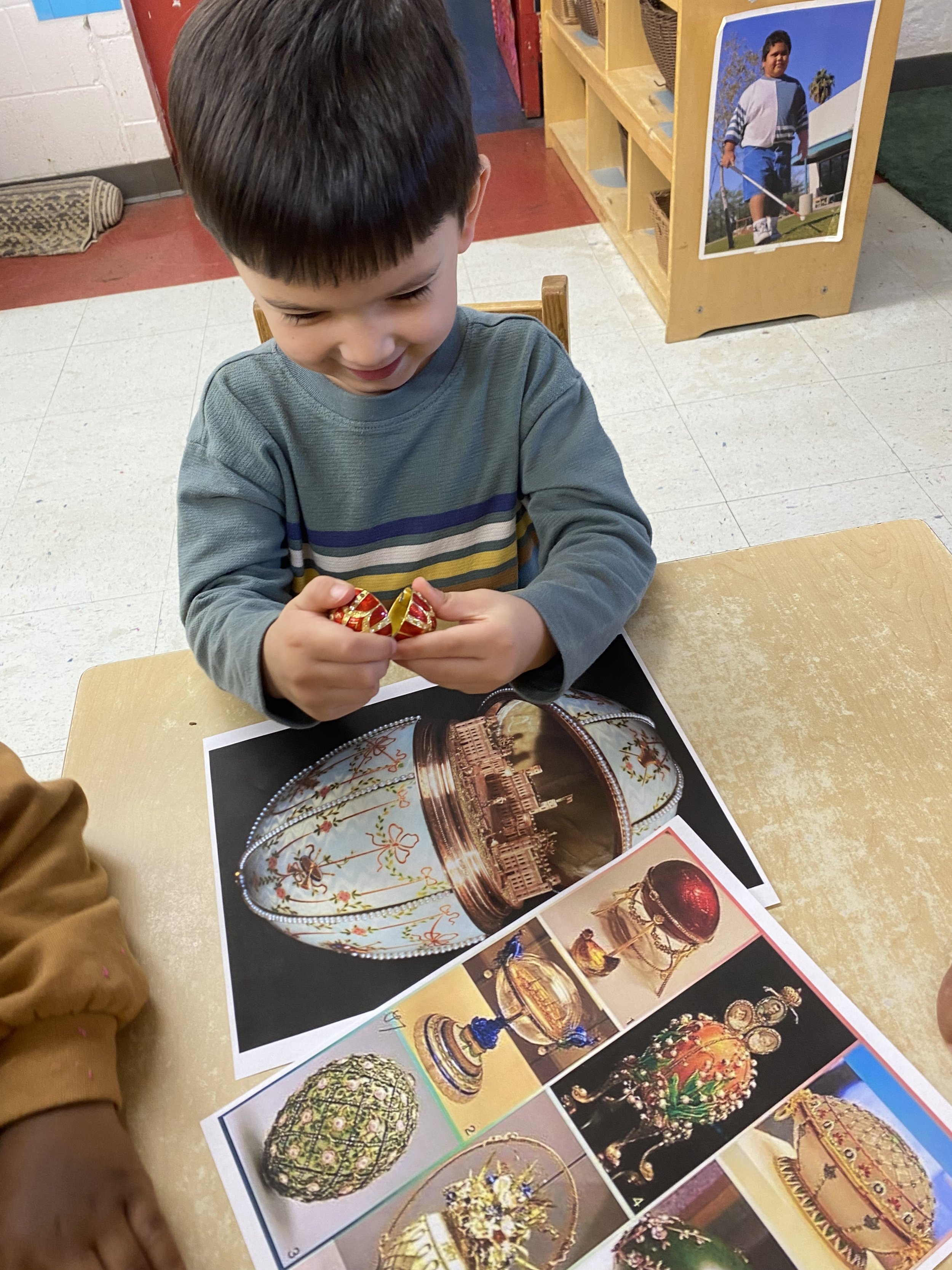 A young boy sitting at a table looking at colorful jewelry, surrounded by pictures of ornate boxes and decorative objects.
