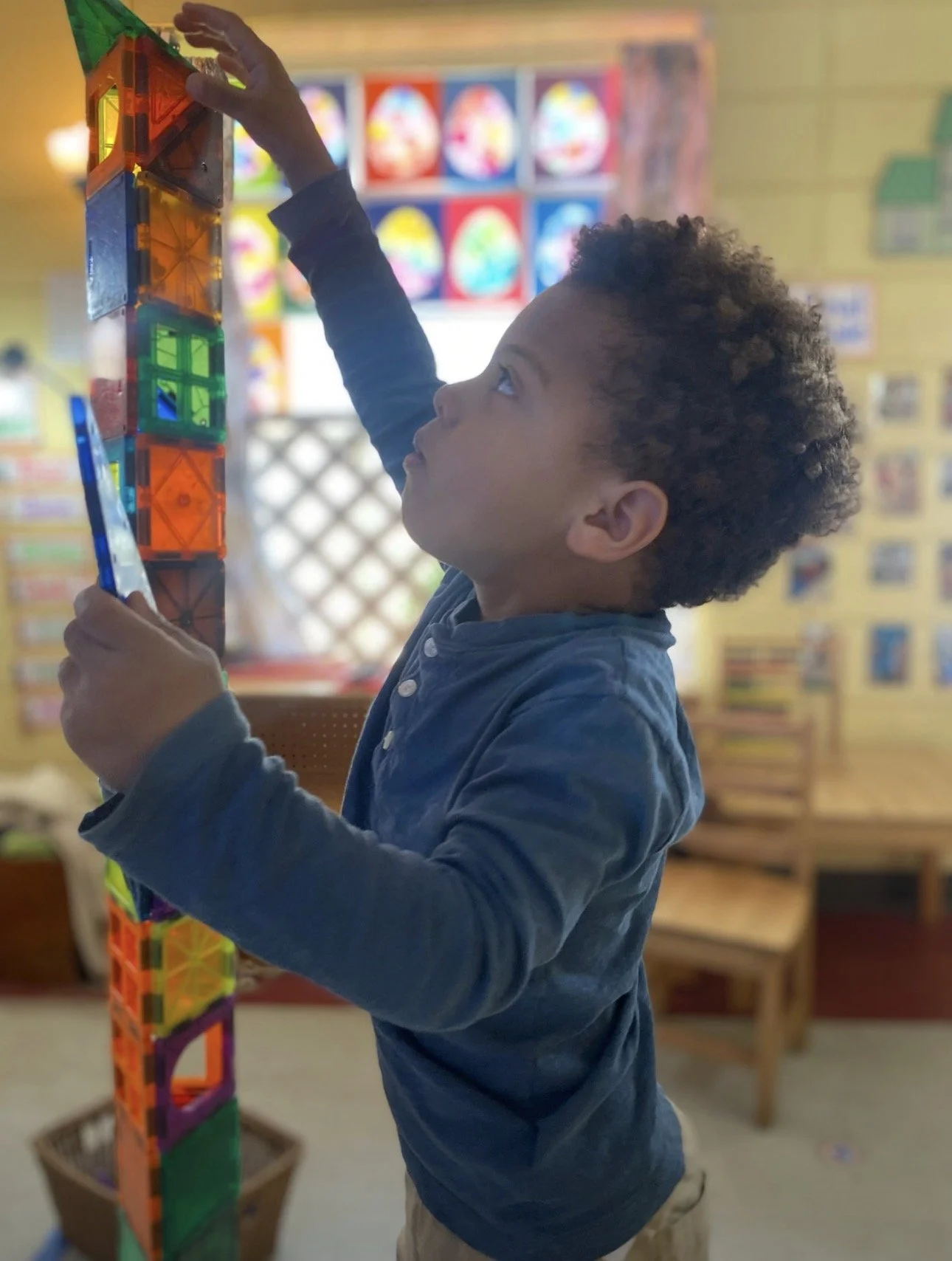 A young boy with curly hair stacking colorful magnetic tiles into a tall structure in a classroom with educational posters in the background.