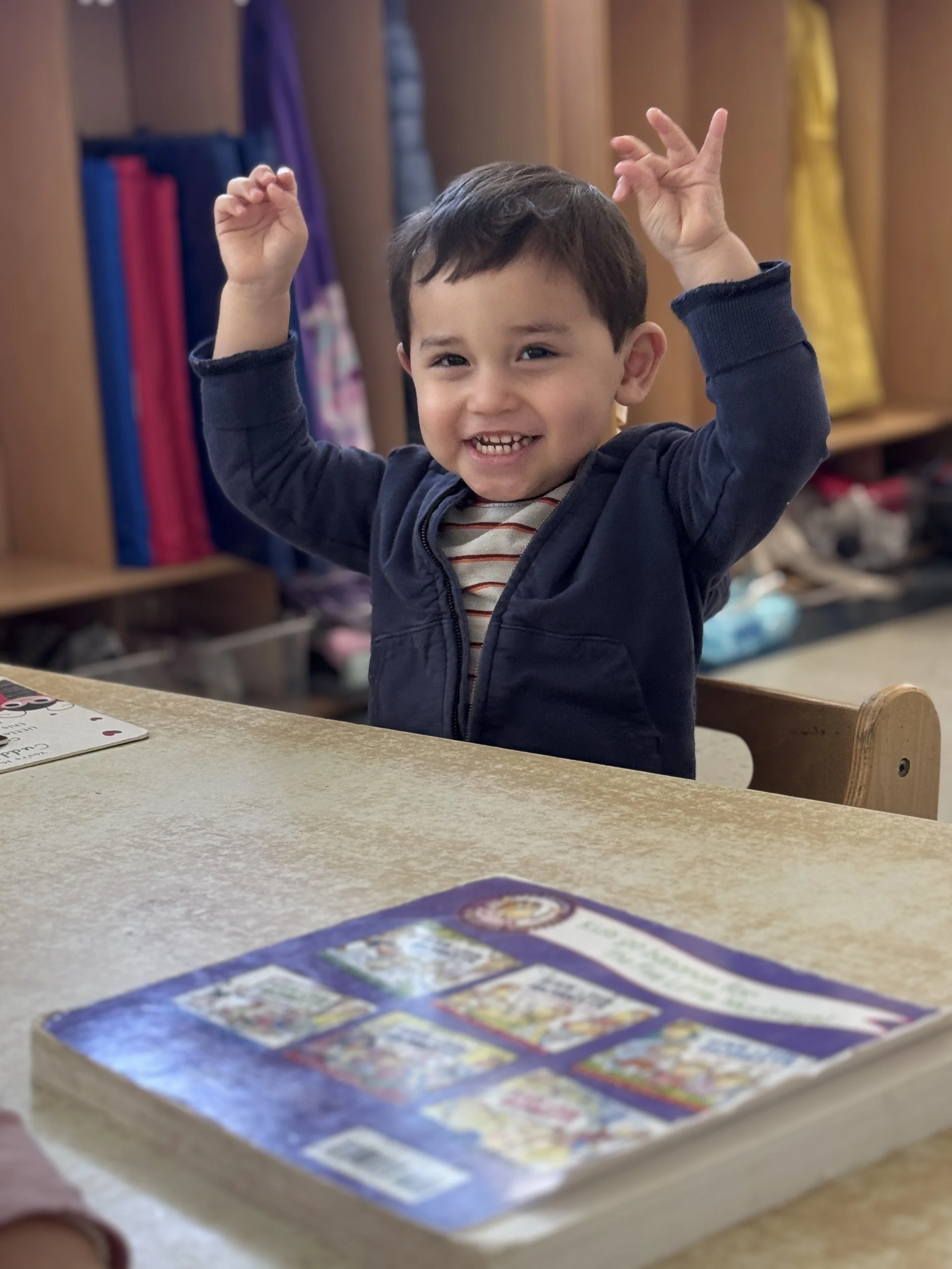 A young boy smiling and raising his arms, sitting at a table with a colorful card game spread out in front of him, in a room with wooden shelves and jackets hanging.