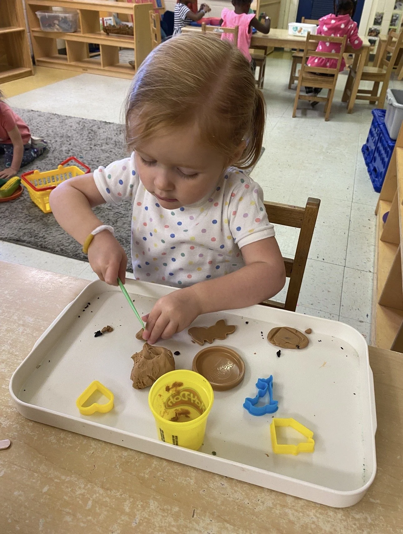 A young girl with red hair wearing a white polka-dot shirt decorates cookies with clay or play dough on a white tray in a classroom.