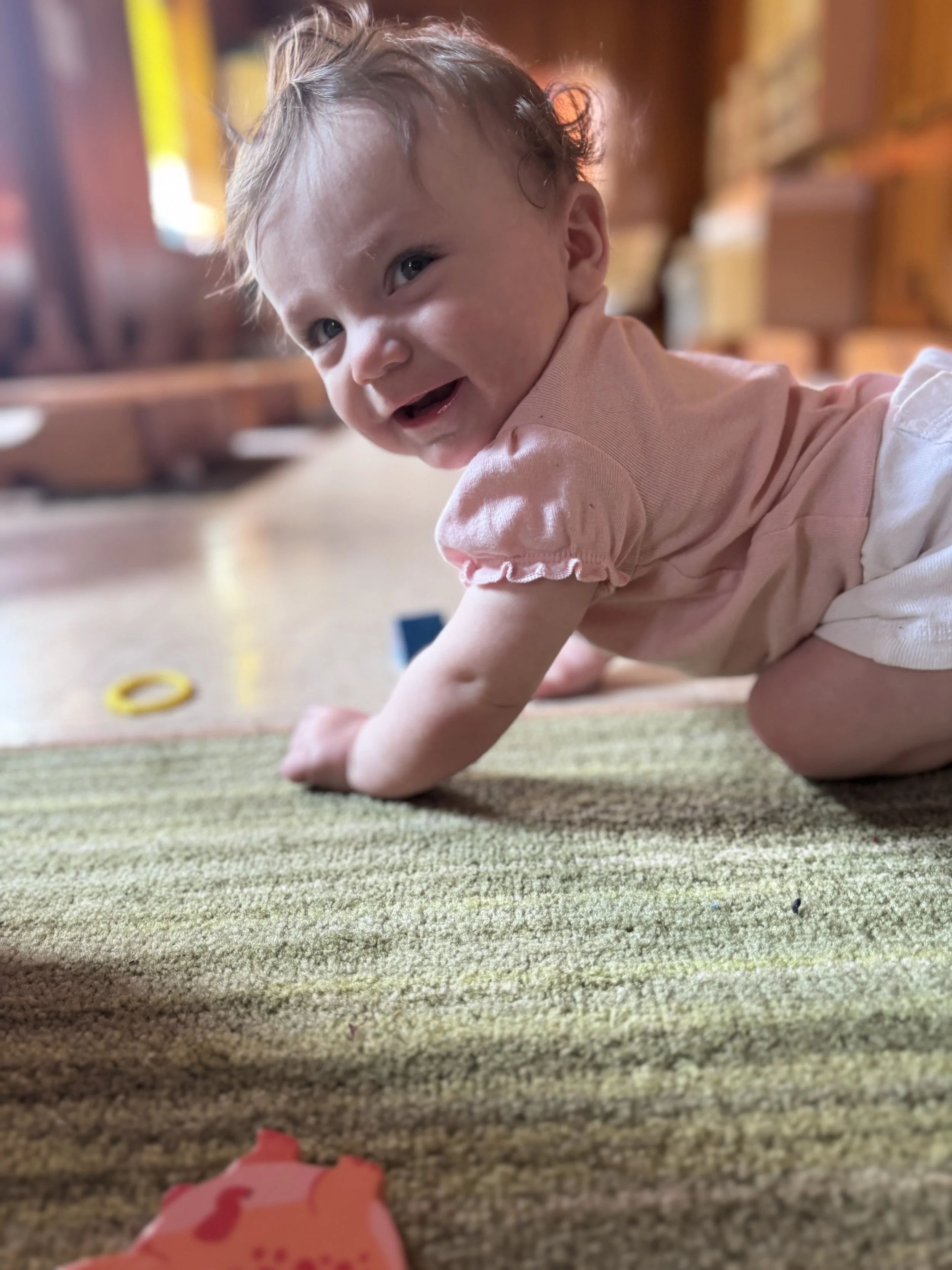 A young child with curly hair smiling and crawling on a carpeted floor indoors, with colorful toys and furniture blurred in the background.