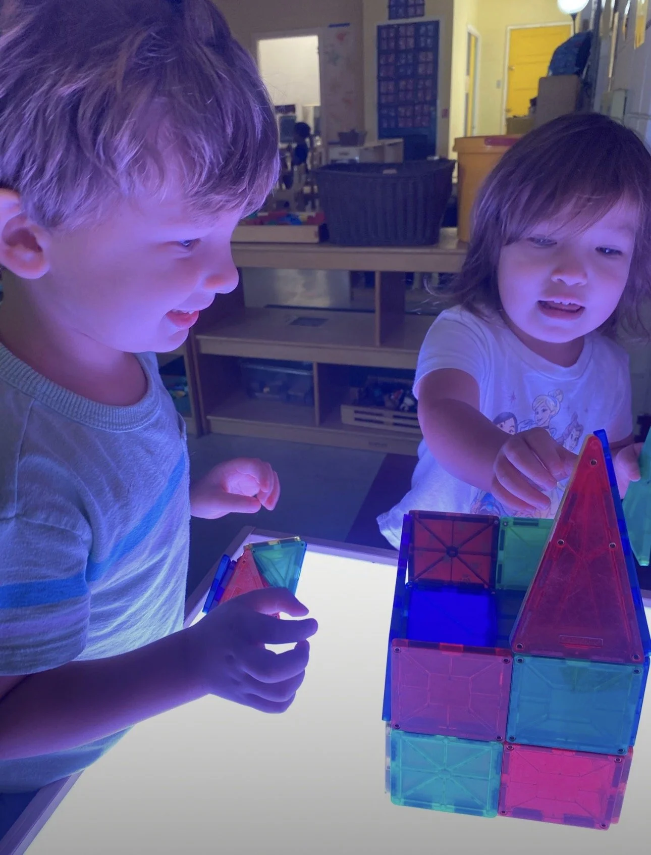 Two children playing with colorful magnetic building blocks on a lighted surface in a classroom.