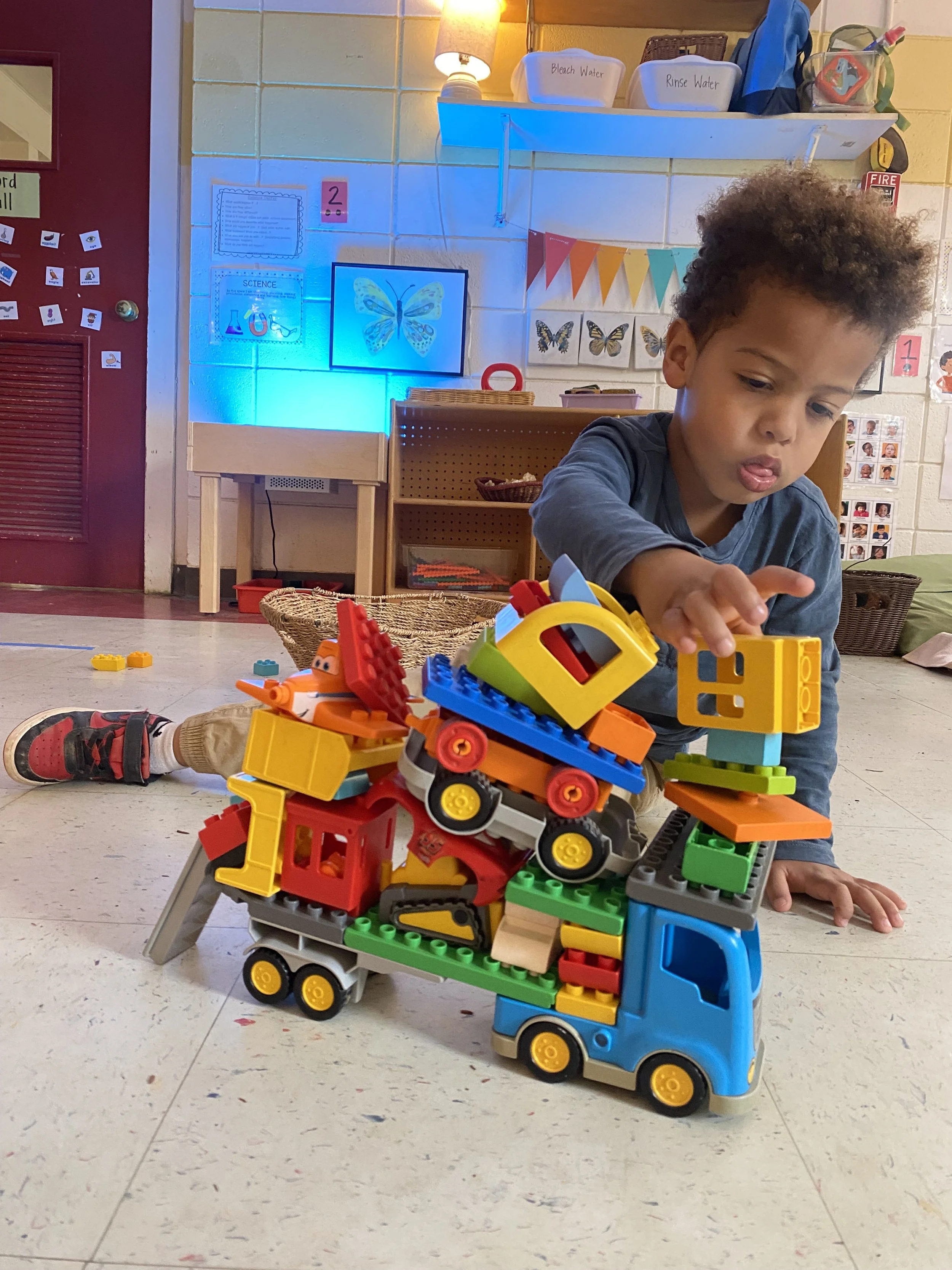 A young boy playing with colorful toy construction vehicles and building blocks on the floor of a classroom or playroom.