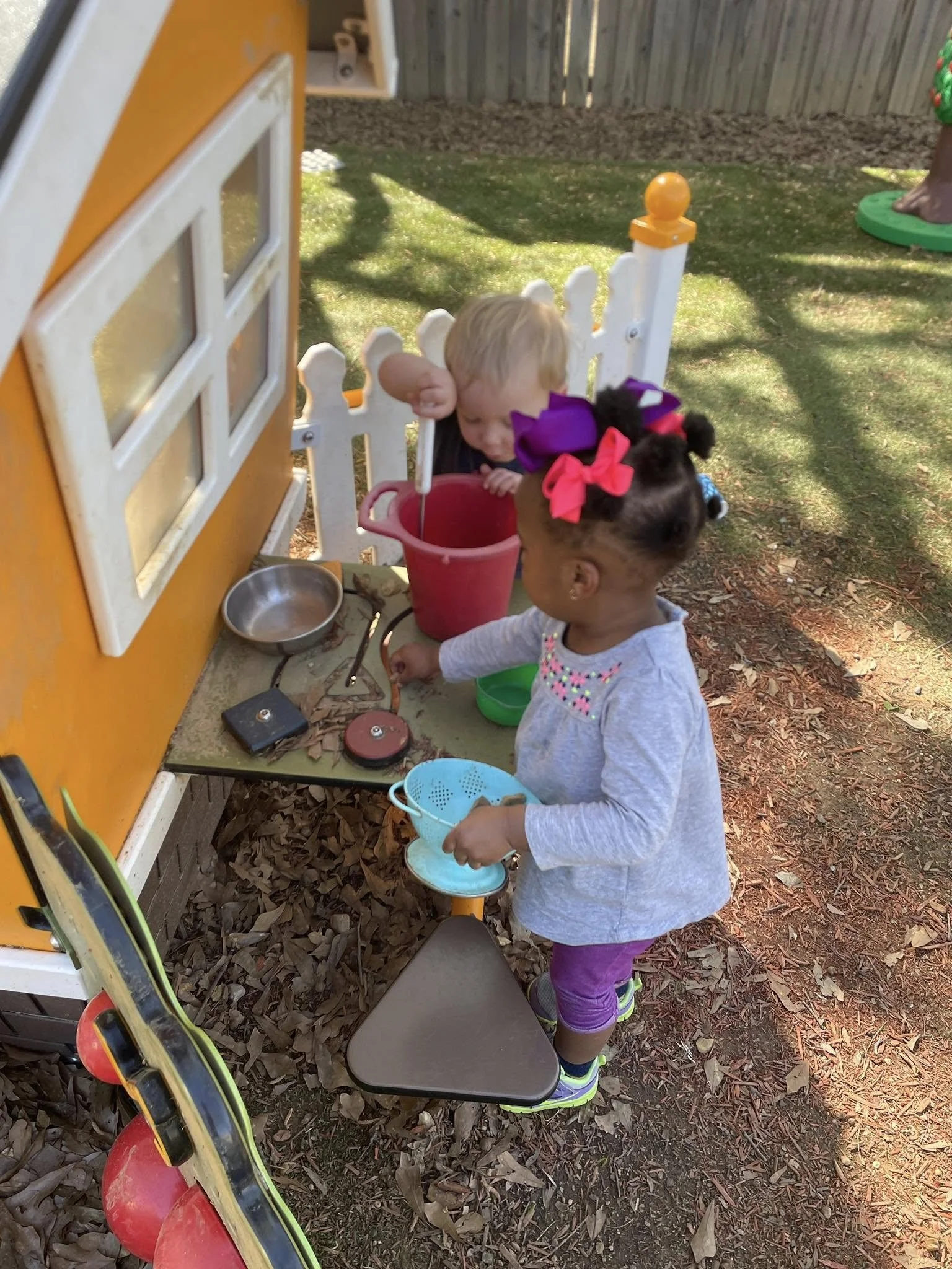 Two young children are playing outside with toy kitchen items, such as a red bucket and a green bowl, near a small playhouse with a yellow exterior. One child is standing on the ground, reaching into a plastic container, while the other is partially visible behind the playhouse. The scene is set on a leaf-covered ground with a wooden fence in the background.