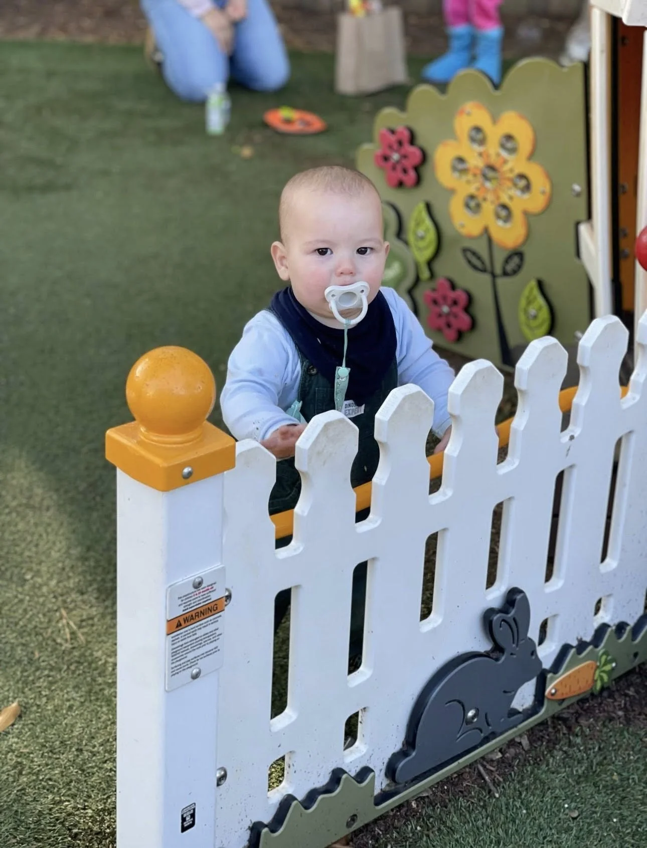 A baby with a pacifier standing behind a small white picket fence at a playground, with colorful flower decorations on a board in the background.