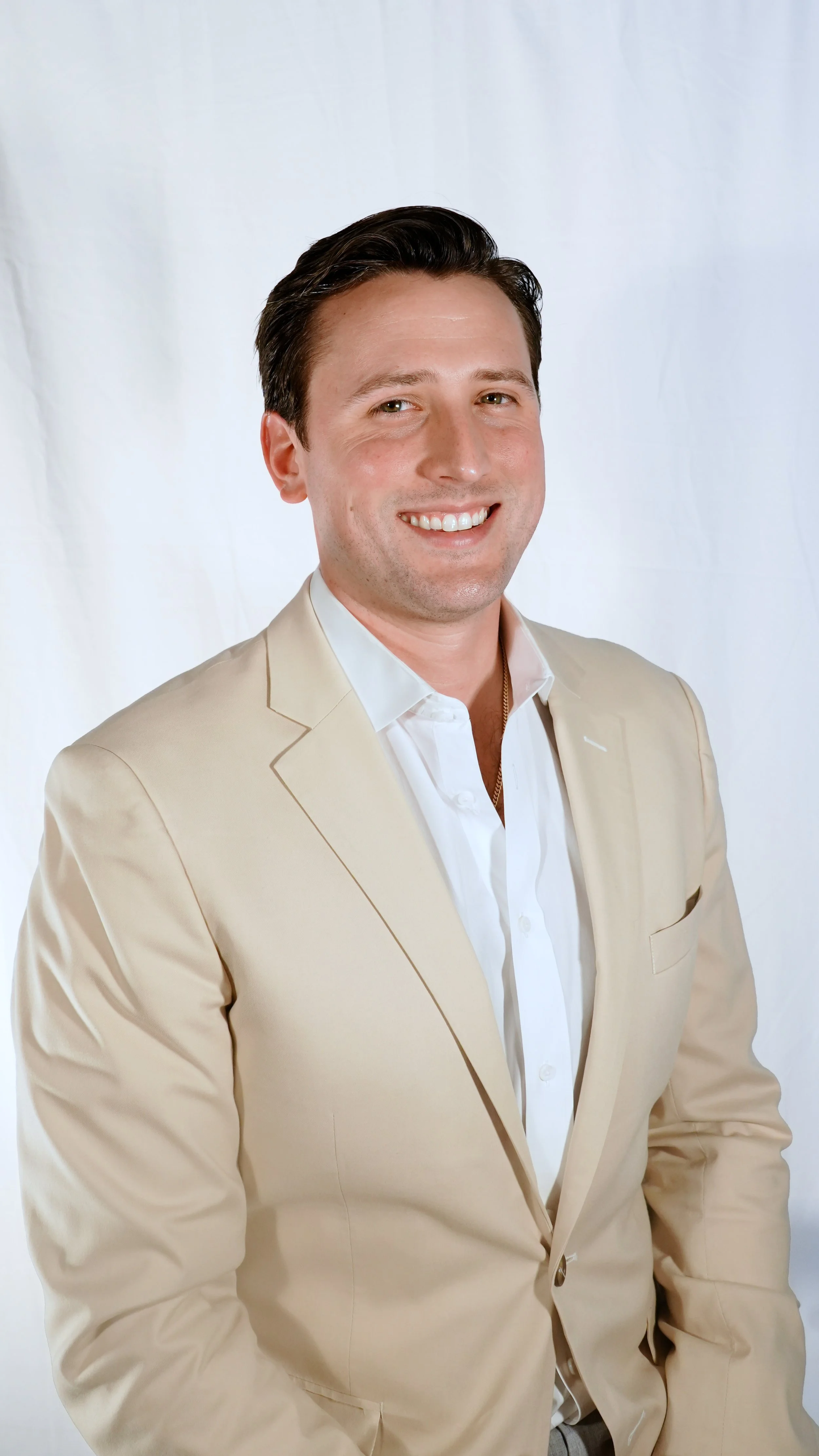A smiling man with dark hair wearing a beige suit jacket and a white shirt against a plain white background.