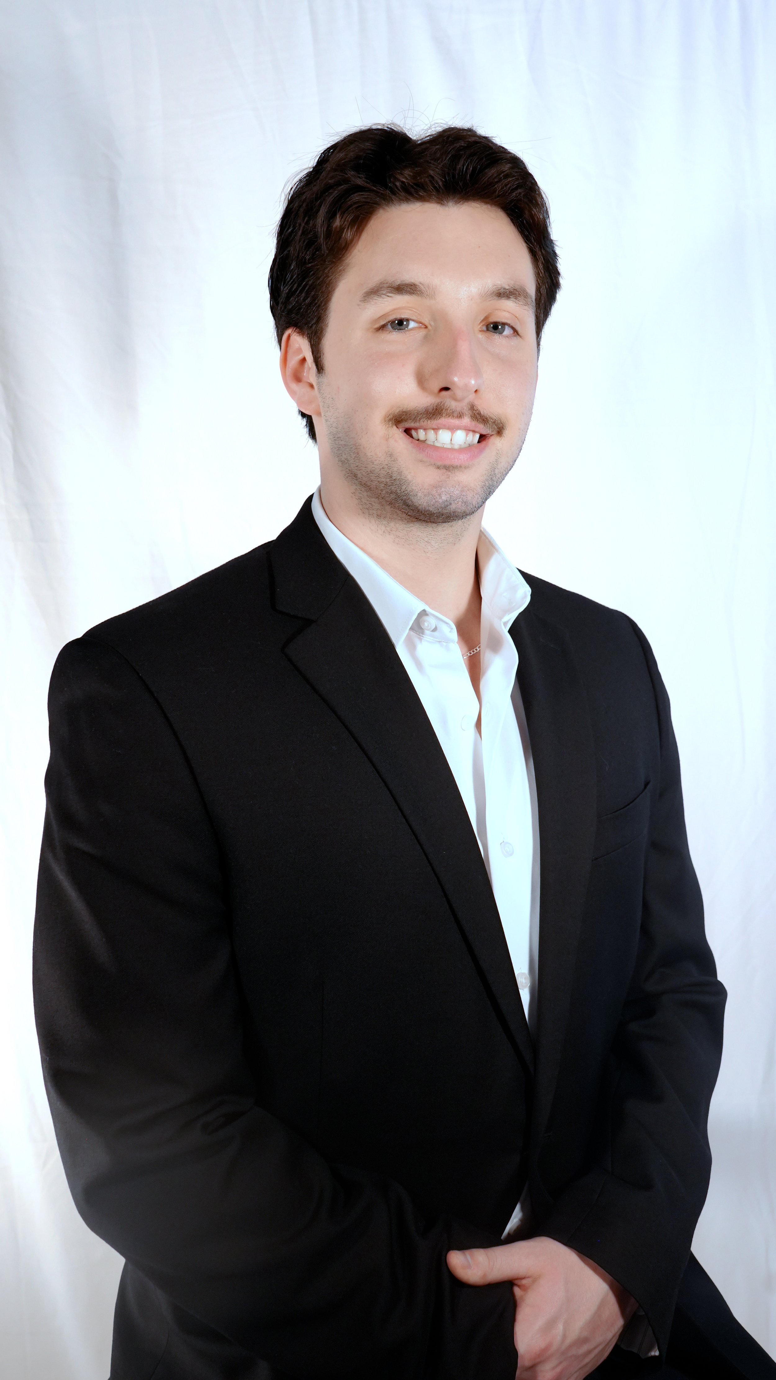 Young man in a black suit and white shirt smiling against a white backdrop.