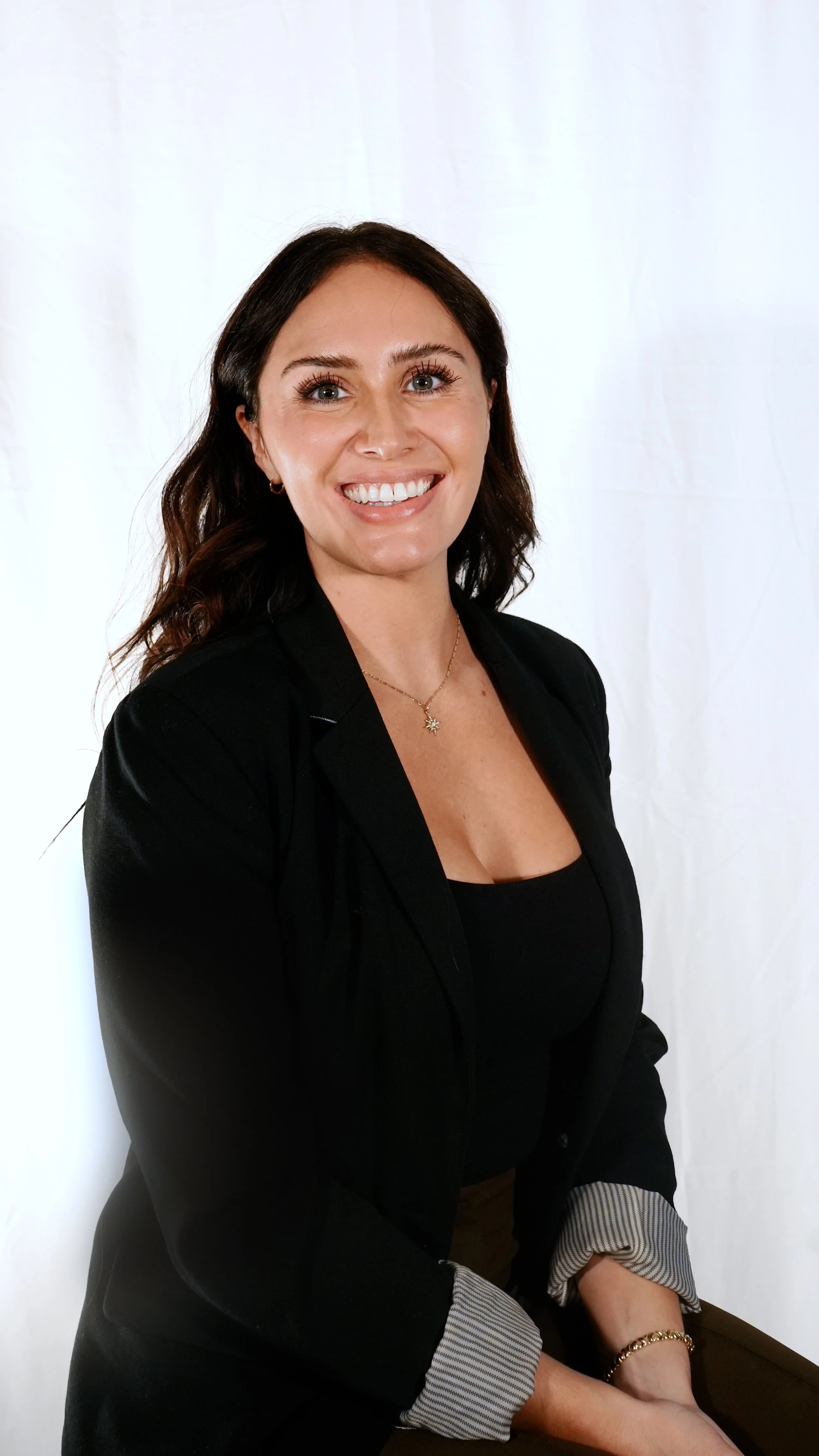 A woman with dark hair wearing a black blazer and shirt, smiling, seated against a white background.