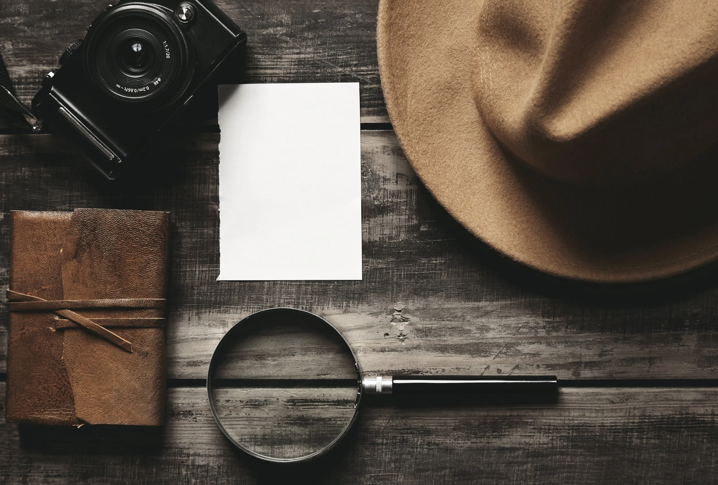 Flat lay of a camera, a leather notebook, a blank white piece of paper, a magnifying glass, and a tan hat on a dark wooden surface.