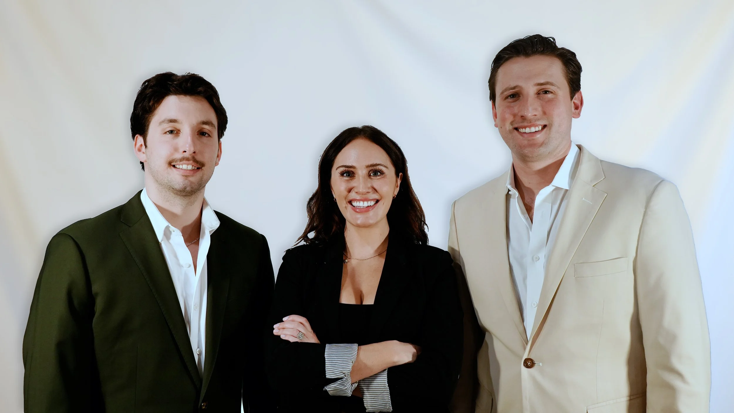 Three smiling professionals standing together against a light background, dressed in business attire.