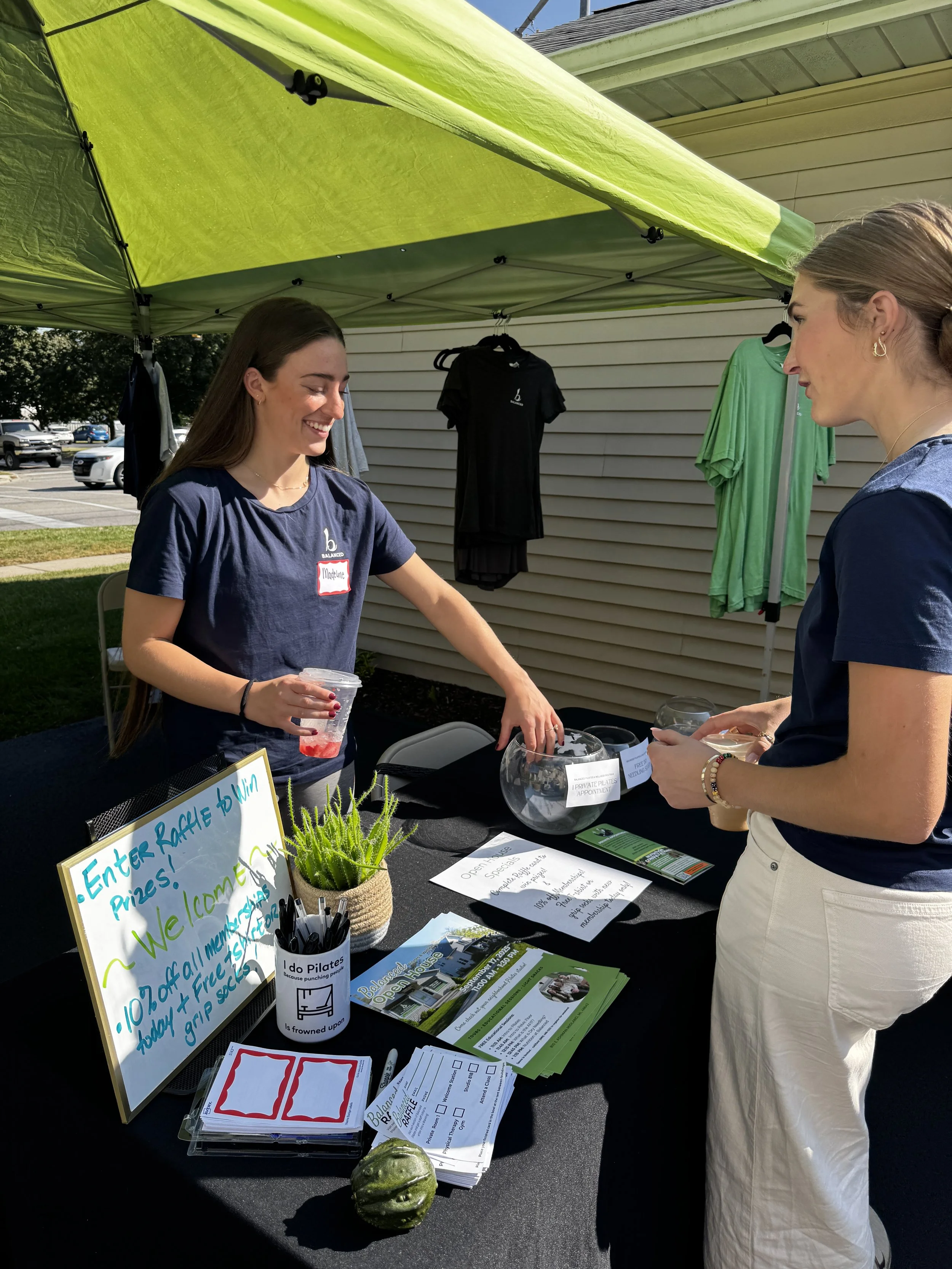 Two women standing at an outdoor booth with informational materials, a whiteboard, and a potted plant, under a green canopy. The woman on the left is smiling and holding a drink, while the woman on the right is engaged in conversation. There are cars