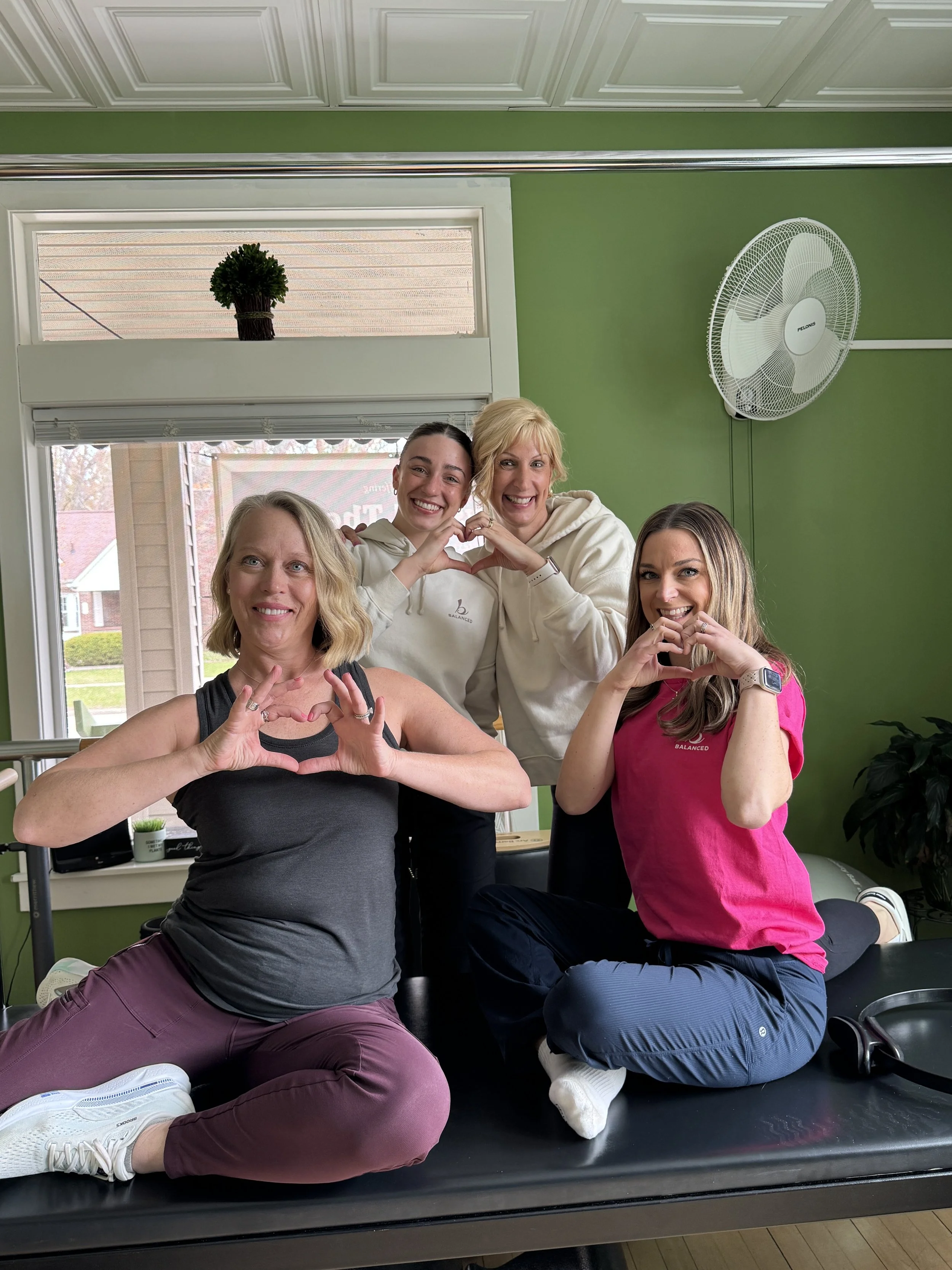 Four women engaging in a fitness or yoga class, sitting on a black mat with two women in the front and two in the back making heart shapes with their hands. They are in a room with green walls, a window, a fan, and a small plant. All are smiling and 