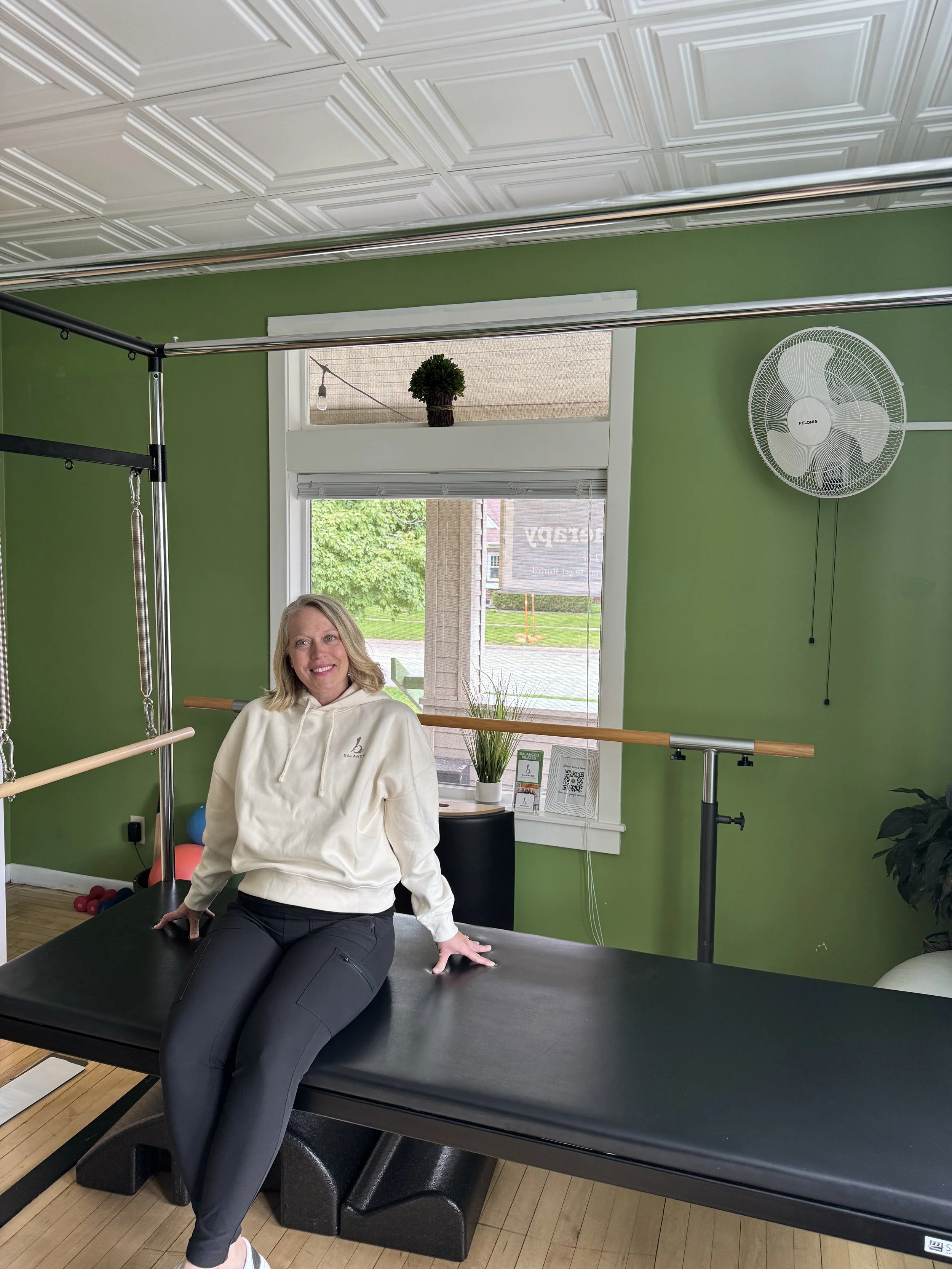 A woman with blonde hair sitting on a Pilates reformer with a black mat in a room with green walls and windows revealing a street and building outside. There is a fan on the wall, and plants near the window.