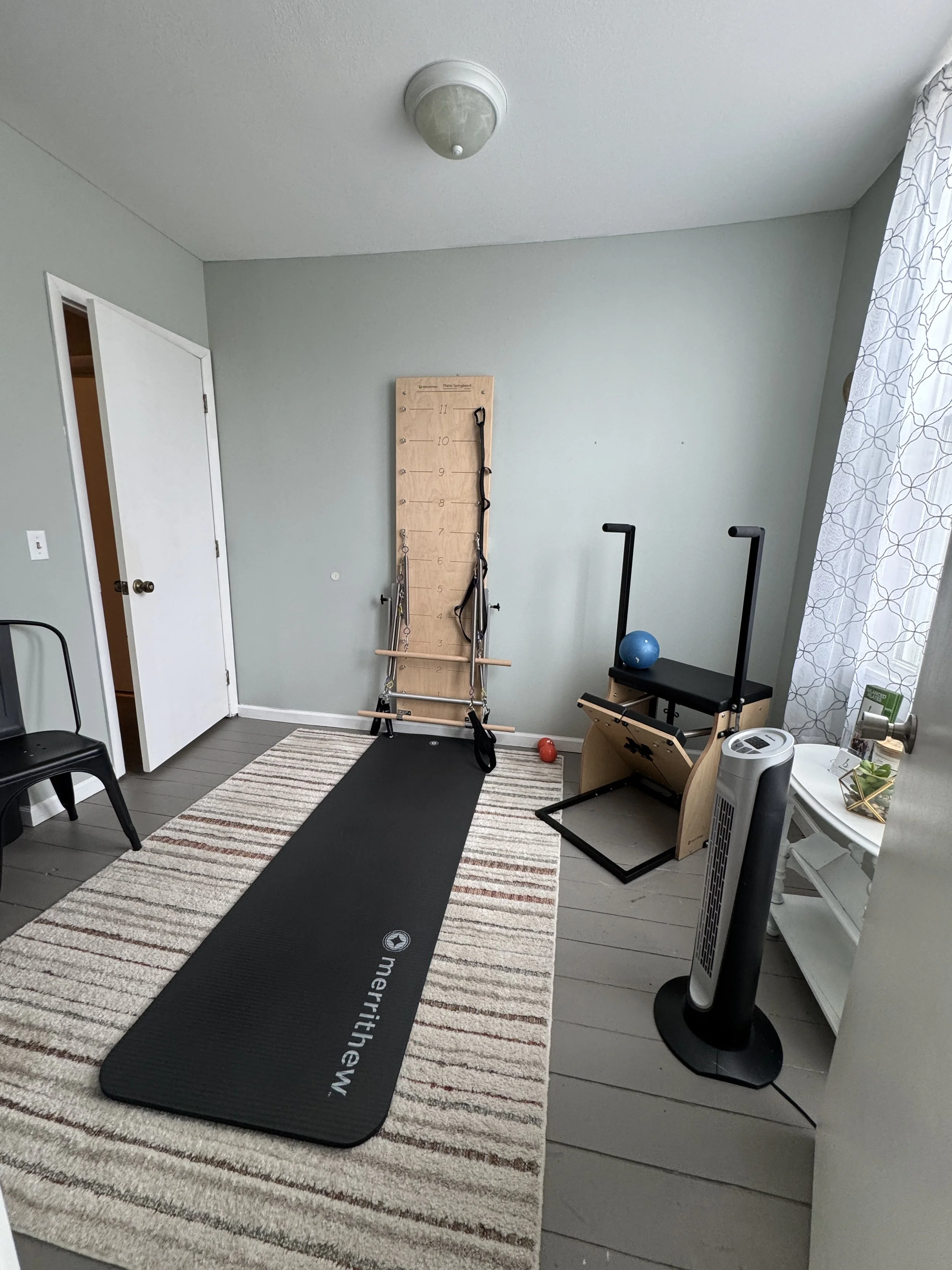 Home gym corner with exercise equipment including a black yoga mat on a striped rug, a wooden wall-mounted organizer with resistance bands and a jump rope, a blue exercise ball, a seated rowing machine, and a standing fan.
