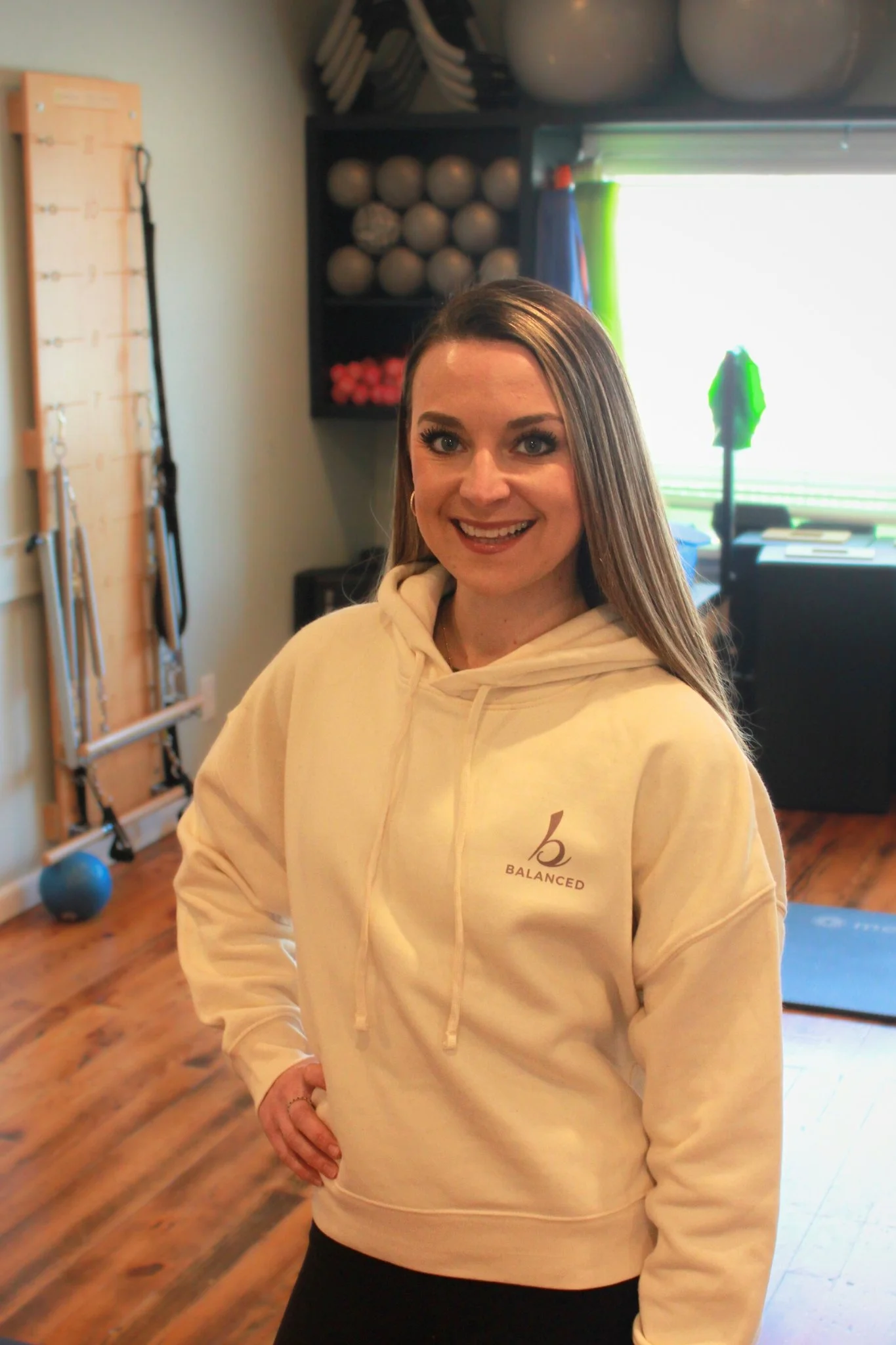 Pilates instructor Rochelle Black wearing a cream-colored hoodie with a logo that says 'Balanced' standing in a fitness studio with workout equipment and a bright window in the background.
