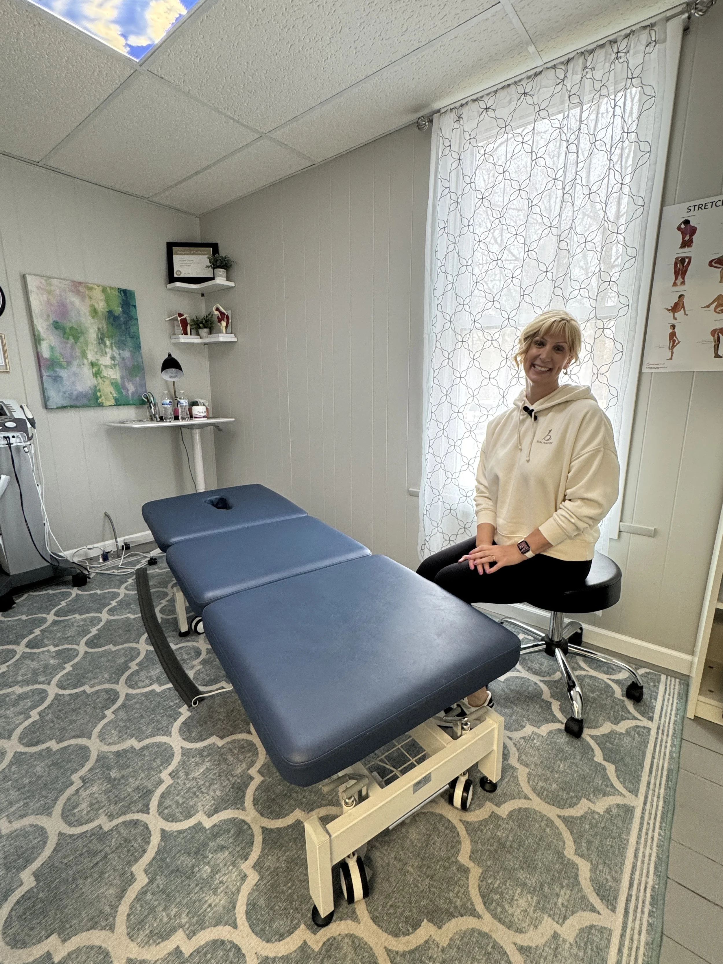 A smiling woman sitting on an examination stool by a physical therapy table in the foreground, medical equipment on the left, and a patterned rug on the floor. 