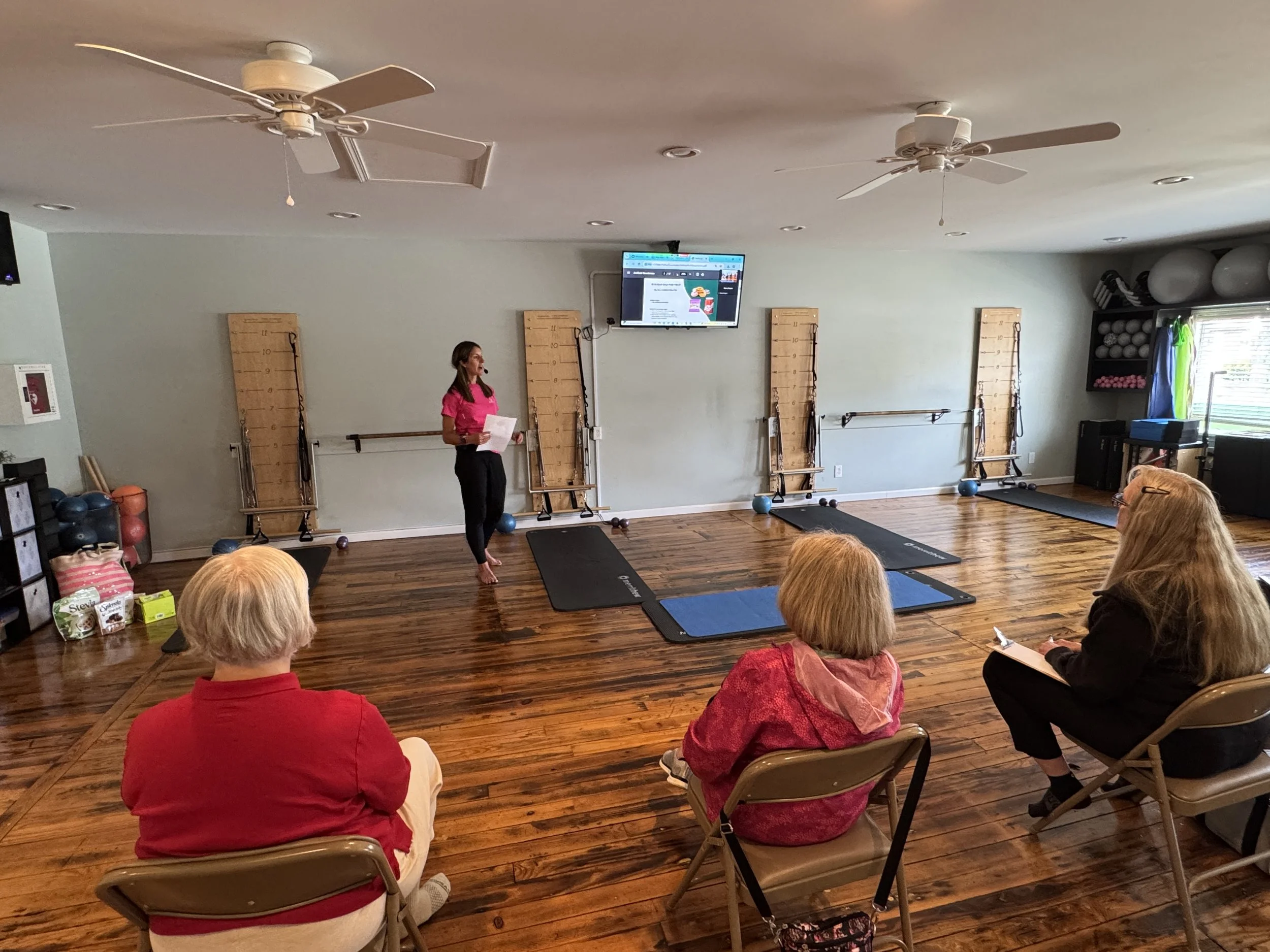 A woman in a pink shirt presents to a small group of people seated in a fitness studio. The room has wooden floors, resistance boards on the wall, and exercise equipment like exercise balls and mats. A TV screen is mounted above, displaying a webpage.