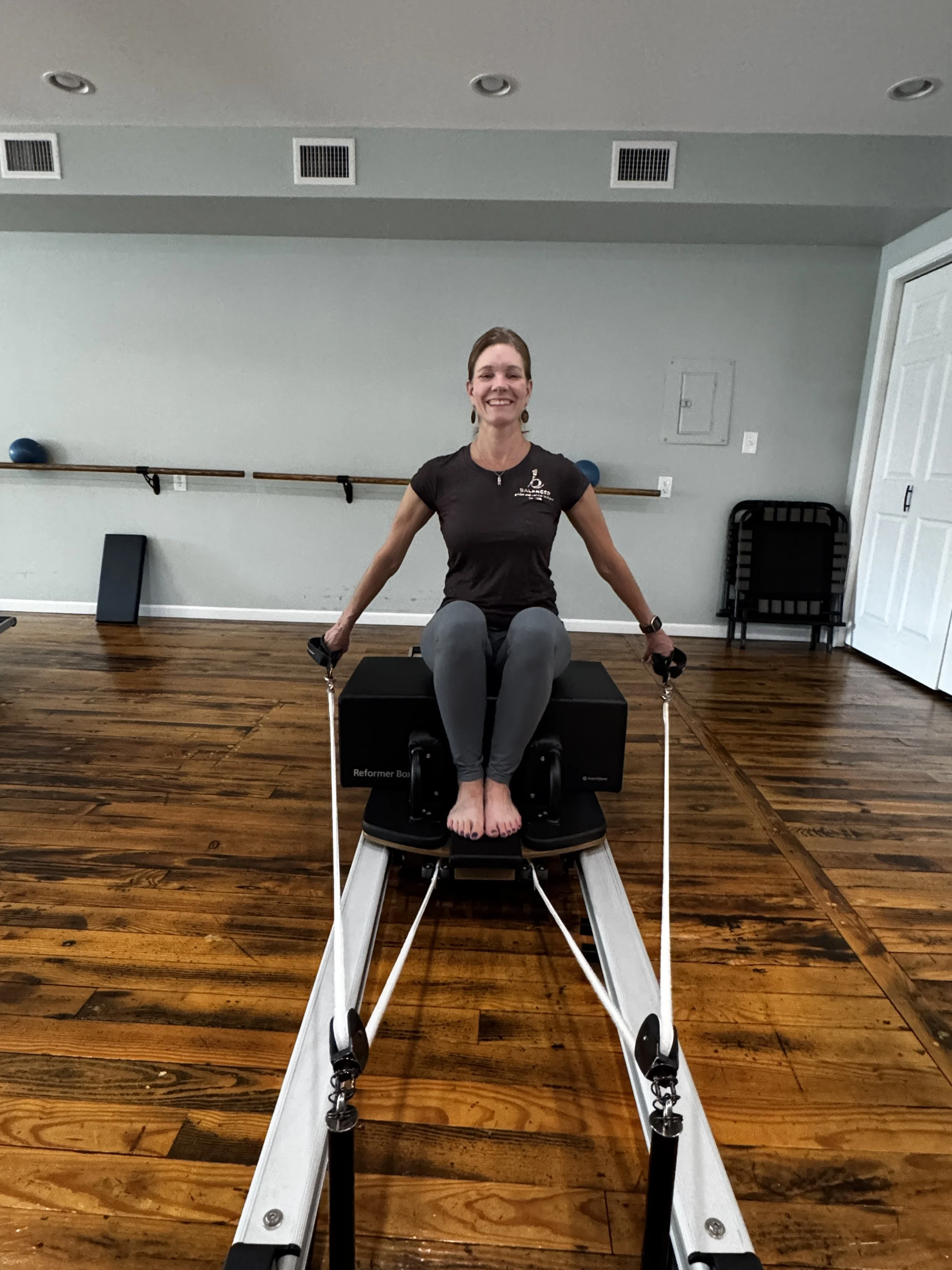 Woman in black t-shirt sitting on a Pilates reformer machine in a Pilates studio in Midland, Michigan smiling at the camera.