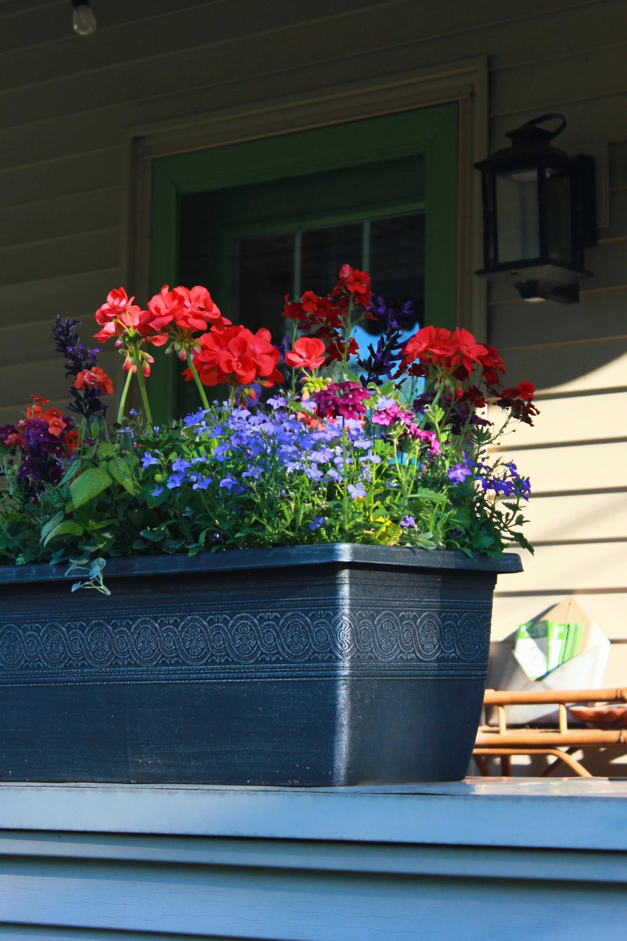 A black flower planter filled with red, purple, and pink flowers on a porch, with a house window and lantern in the background.