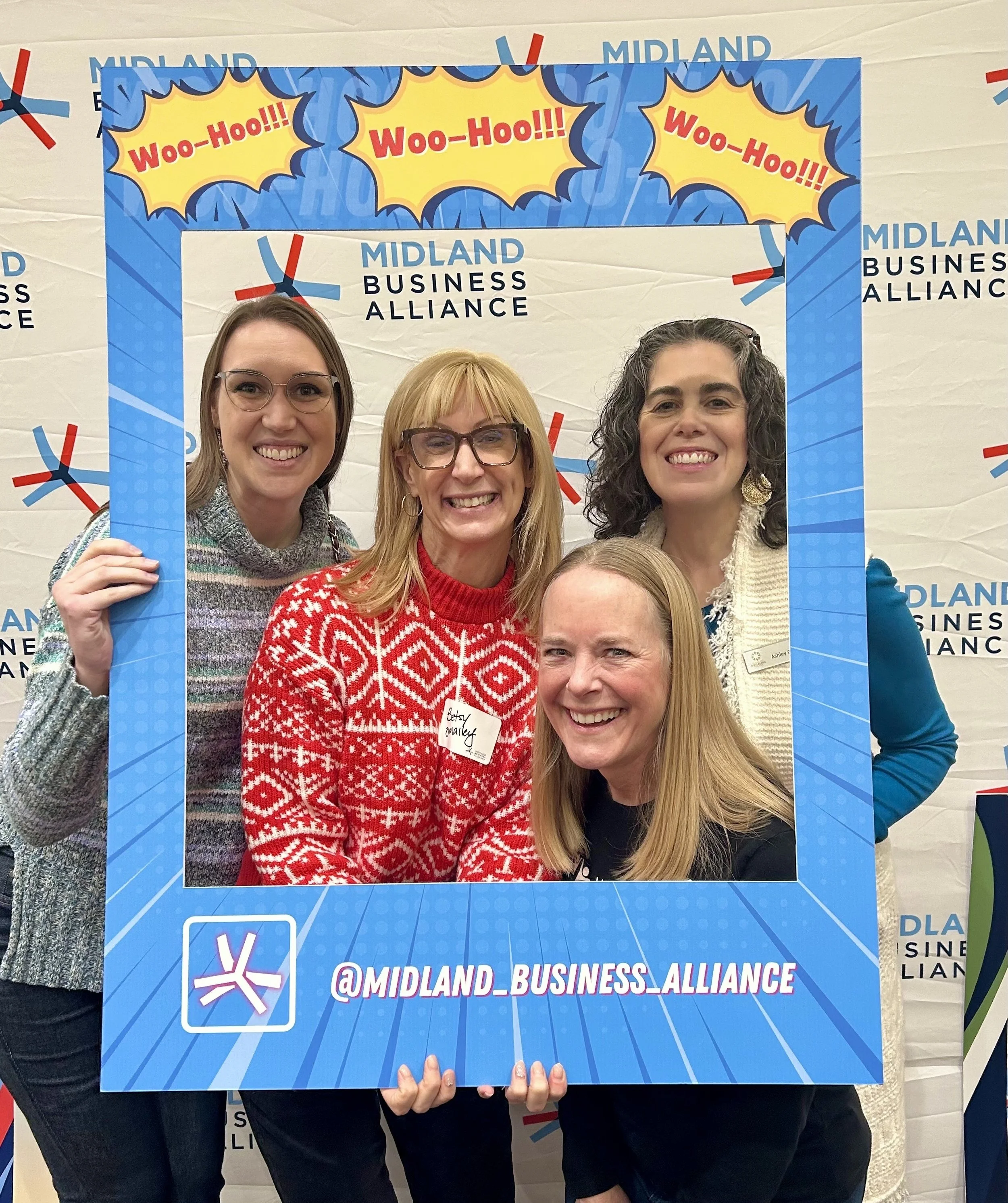 Four women of Balanced Pilates and Wellness Boutique and the Midland Area Wellbeing Coalition at a Midland Business Alliance event holding a large blue photo frame with 'Woo-Hoo!!!'