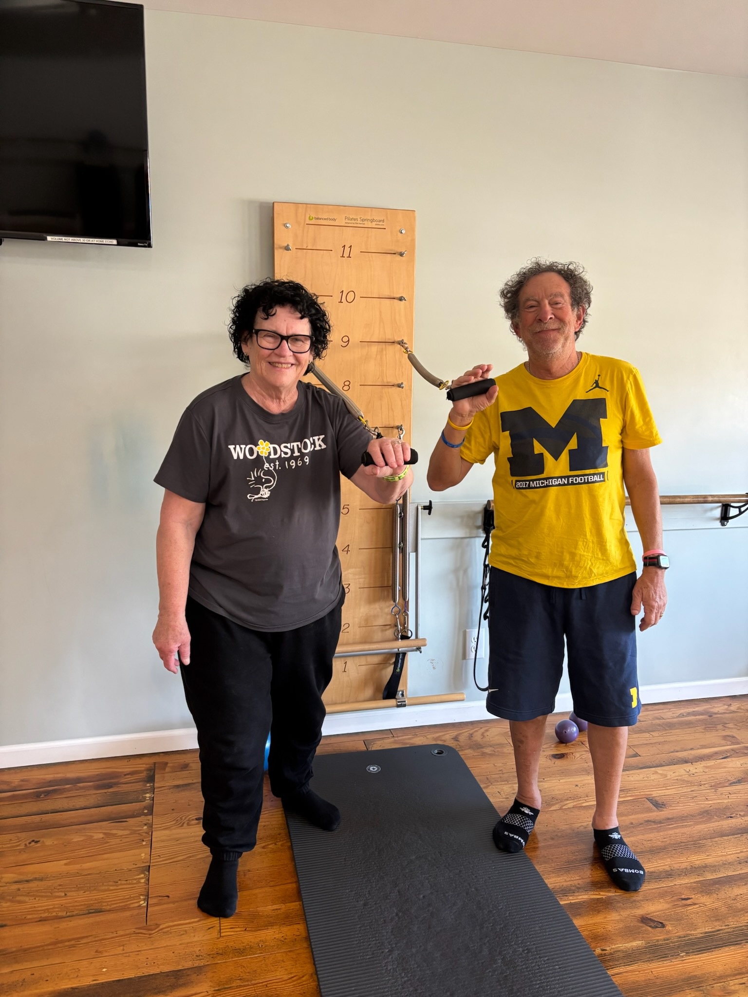 Two people standing indoors, holding gray resistance bands at Balanced Pilates and Wellness Boutique in Midland, Michigan. They are next to a wooden height measurement board and on a black exercise mat, with a wooden floor.