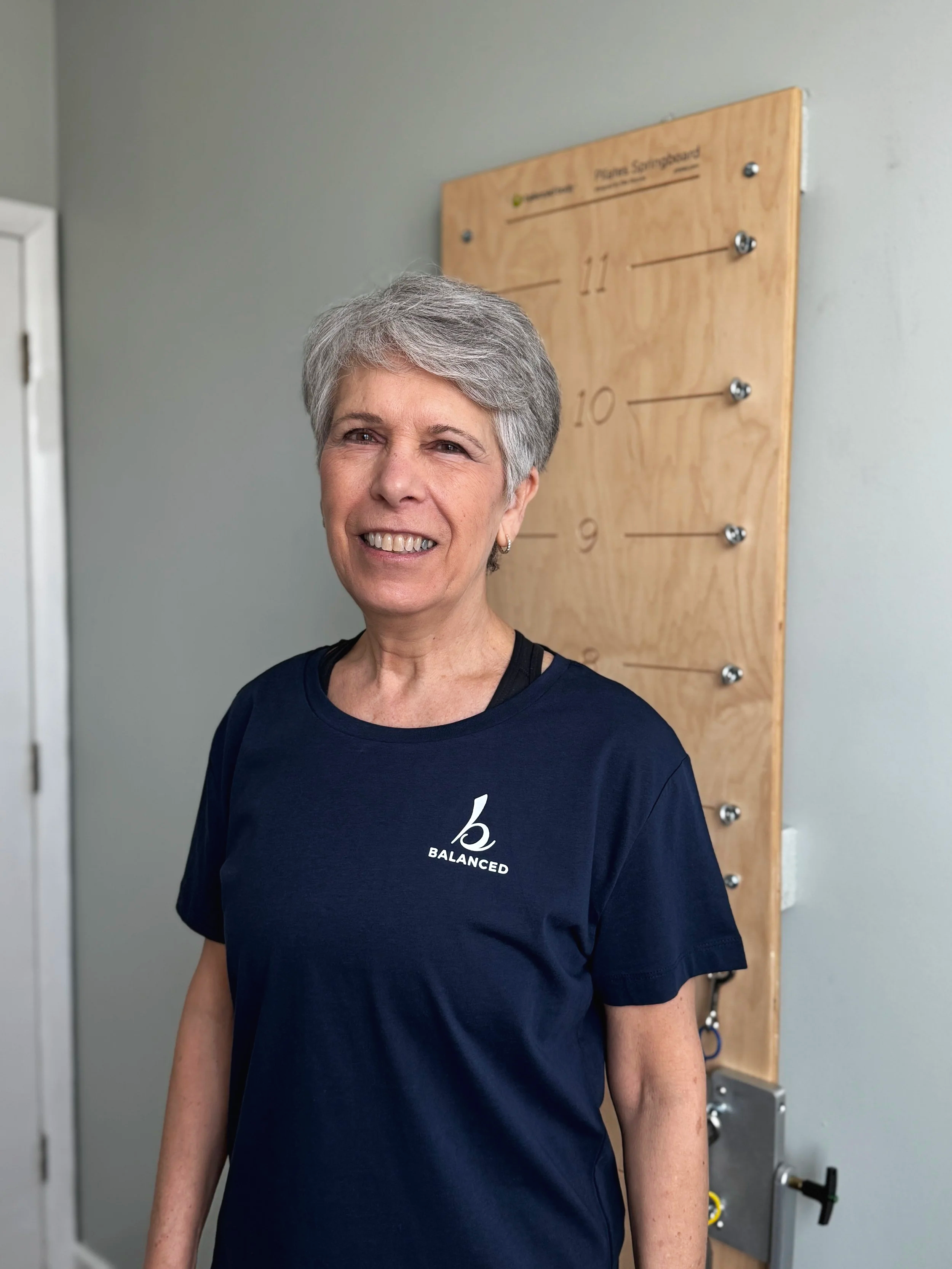 Pilates instructor Judi Powers with short gray hair wearing a navy blue t-shirt with a meditation logo and the word "BALANCED" on it, standing in front of a wall-mounted wooden pegboard in a fitness or therapy center.