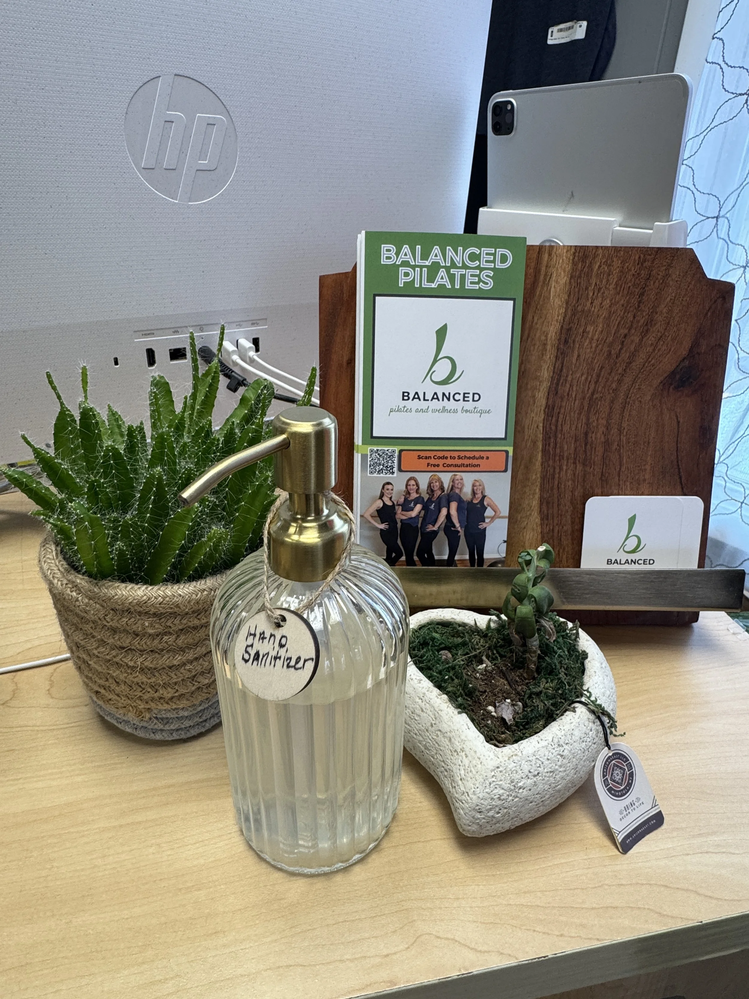 A display of various items on a wooden table including a potted cactus, a small plant in a soil-filled white textured pot, a hand sanitizer bottle, and a brochure for 'Balanced Pilates'. In the background, there are wooden file holders and a portable