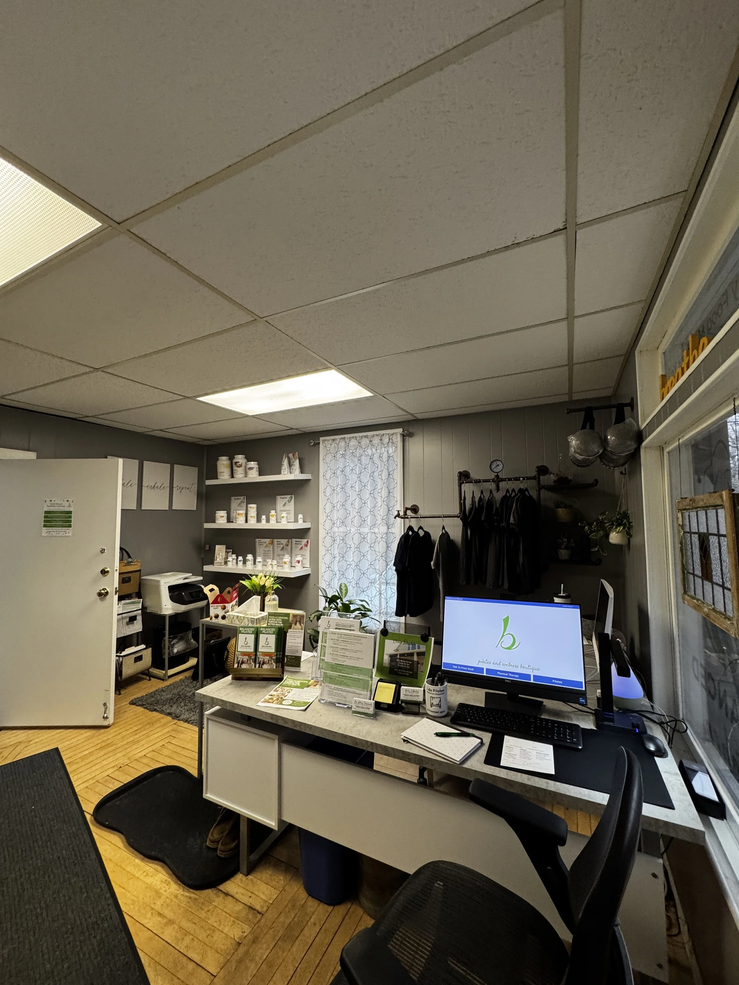 Interior of a wellness boutique and physical therapy office with a reception desk, computer, and display brochures, with shelves of supplement jars and clothing racks at Balanced Pilates and Wellness Boutique in Midland, Michigan.