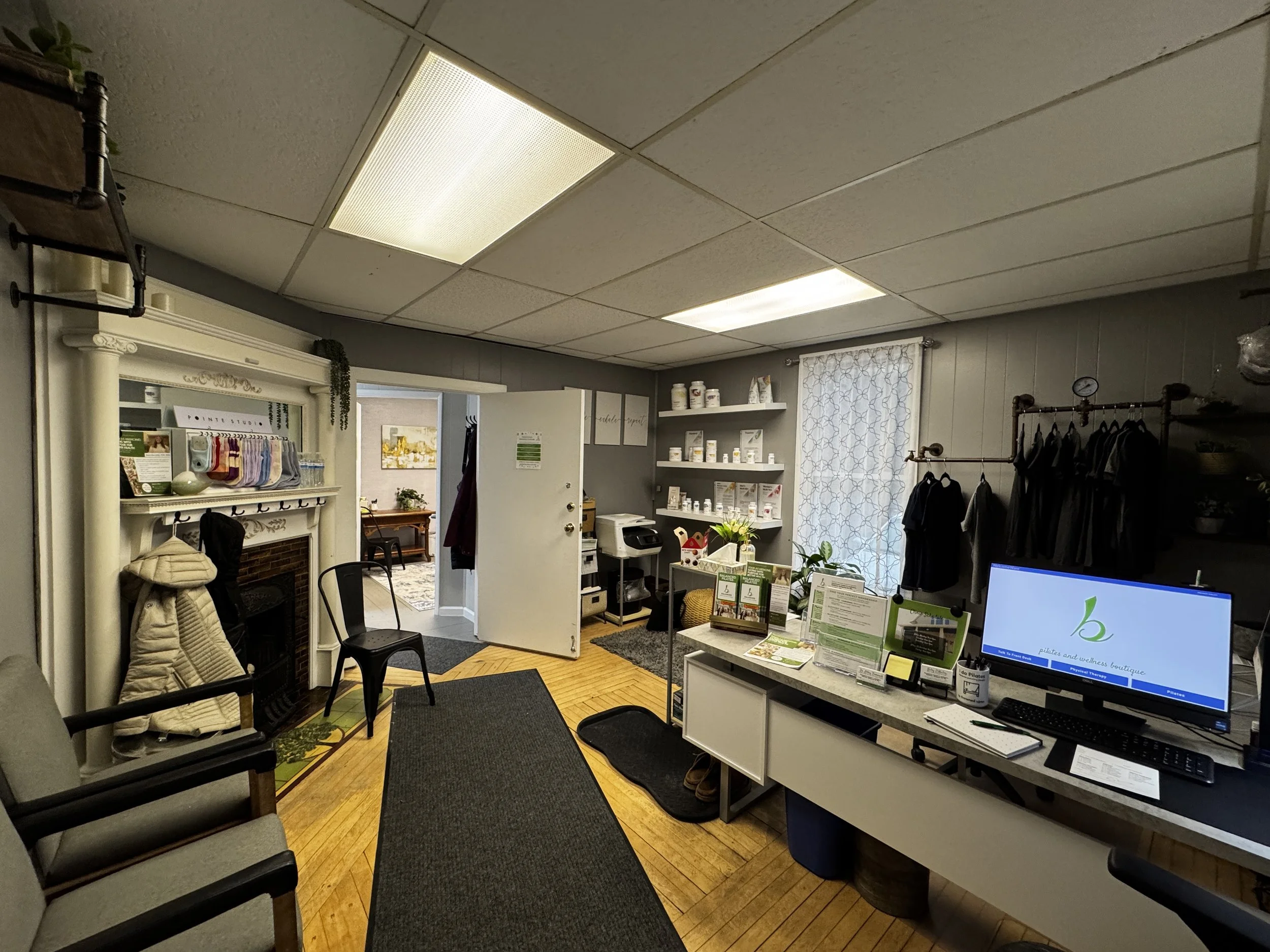 Interior of a wellness and health boutique featuring a reception desk with a computer, shelves with supplements, a coat rack, and a comfy seating area with chairs and a decorative fireplace at Balanced Pilates and Wellness Boutique in Midland.