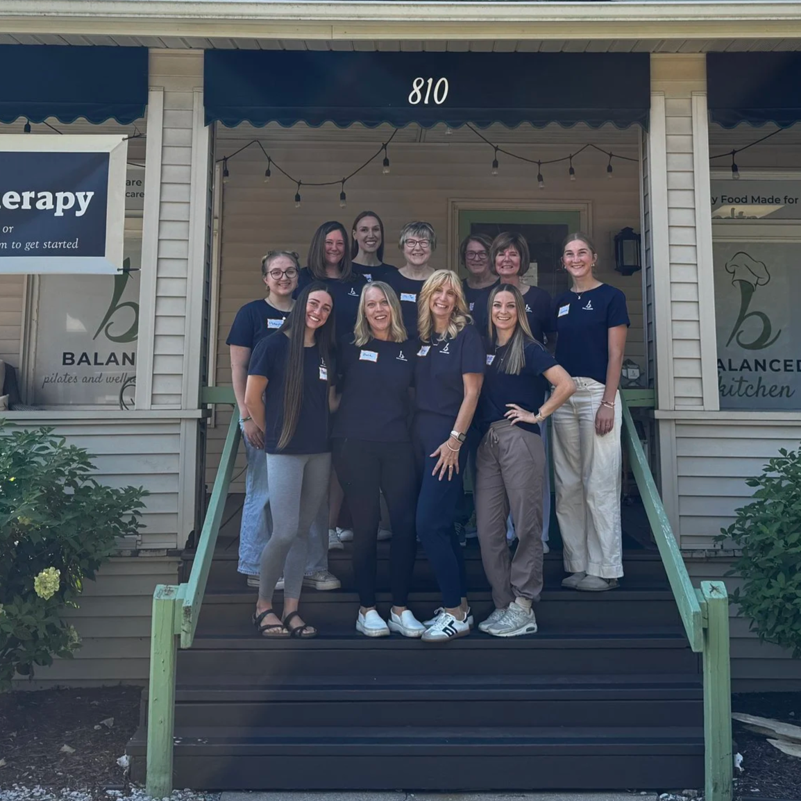 Group of eleven women standing on a porch in front of Balanced Pilates and Wellness Boutique in Midland, Michigan. They are dressed in matching navy blue shirts, smiling, and posing for the photo.