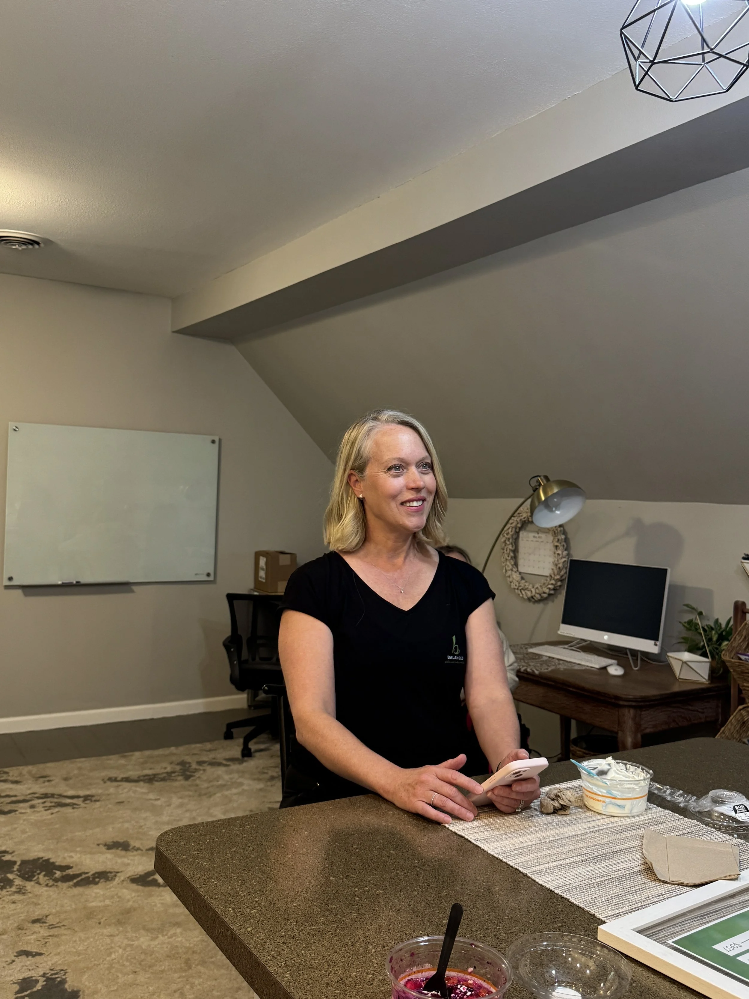 A woman sitting at a table, holding a smartphone, with dessert and food containers on the table. In the background, there is a whiteboard, computer, and desk lamp in an office setting.