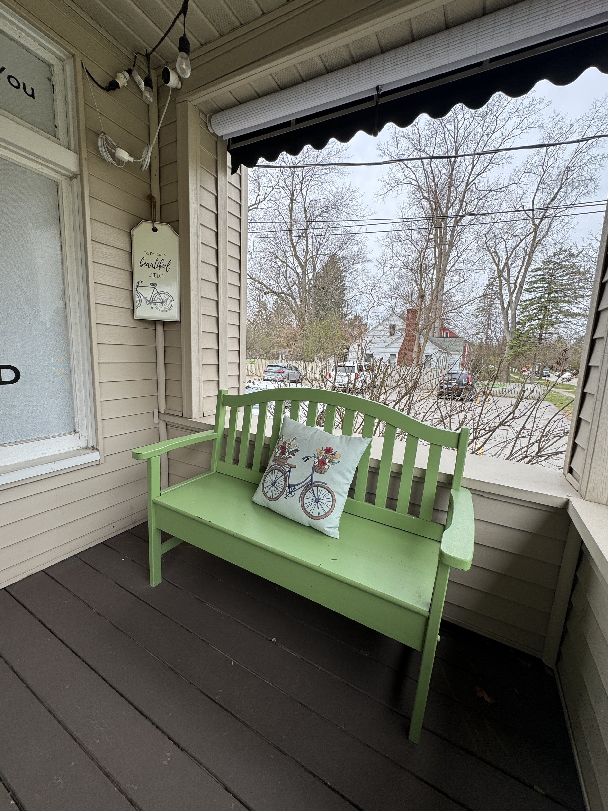 A green wooden bench with a cushions featuring a bicycle with a basket of flowers, on a porch with brown wooden floorboards, beige siding, and a view of trees and houses in the background.