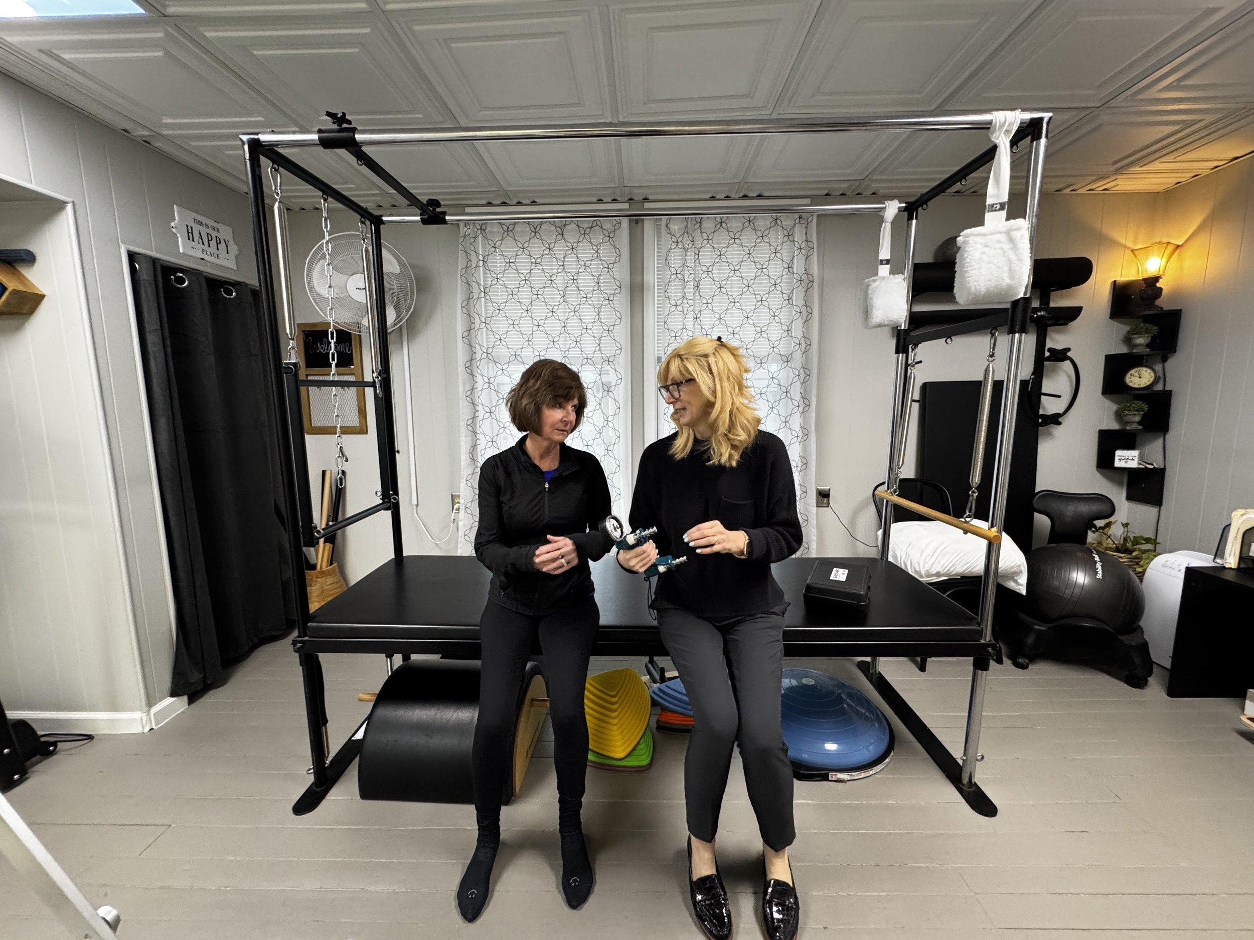 Two women standing in private physical therapy gym with exercise equipment, and workout accessories, engaging in conversation at Balanced Pilates and Wellness Boutique in Midland, Michigan.