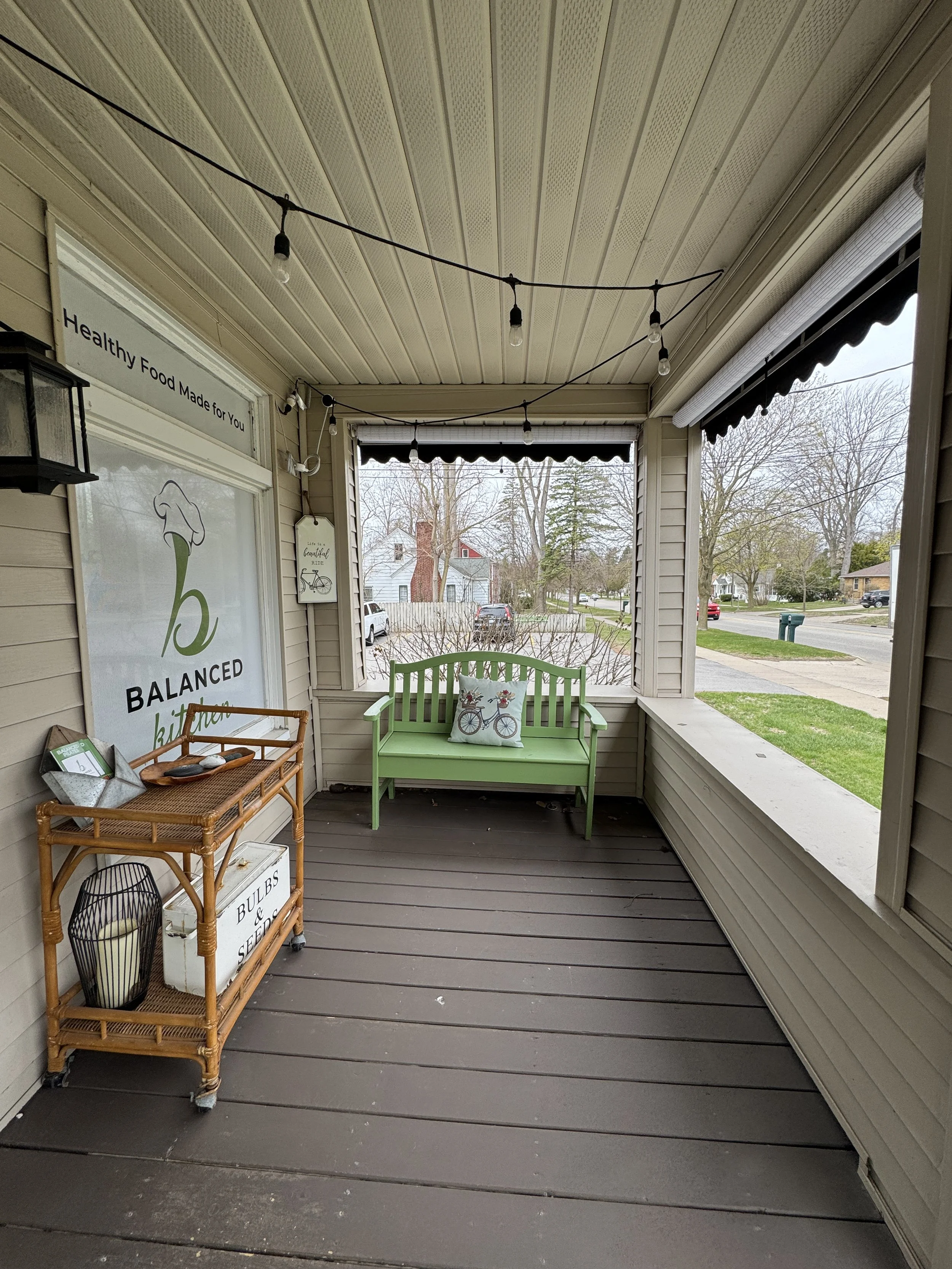 View of a cozy porch with a light green bench with a cushion depicting a bicycle, a wicker cart with souvenirs including a sign and a wire basket, string lights hanging from the ceiling, and a view of a residential neighborhood with trees and houses.