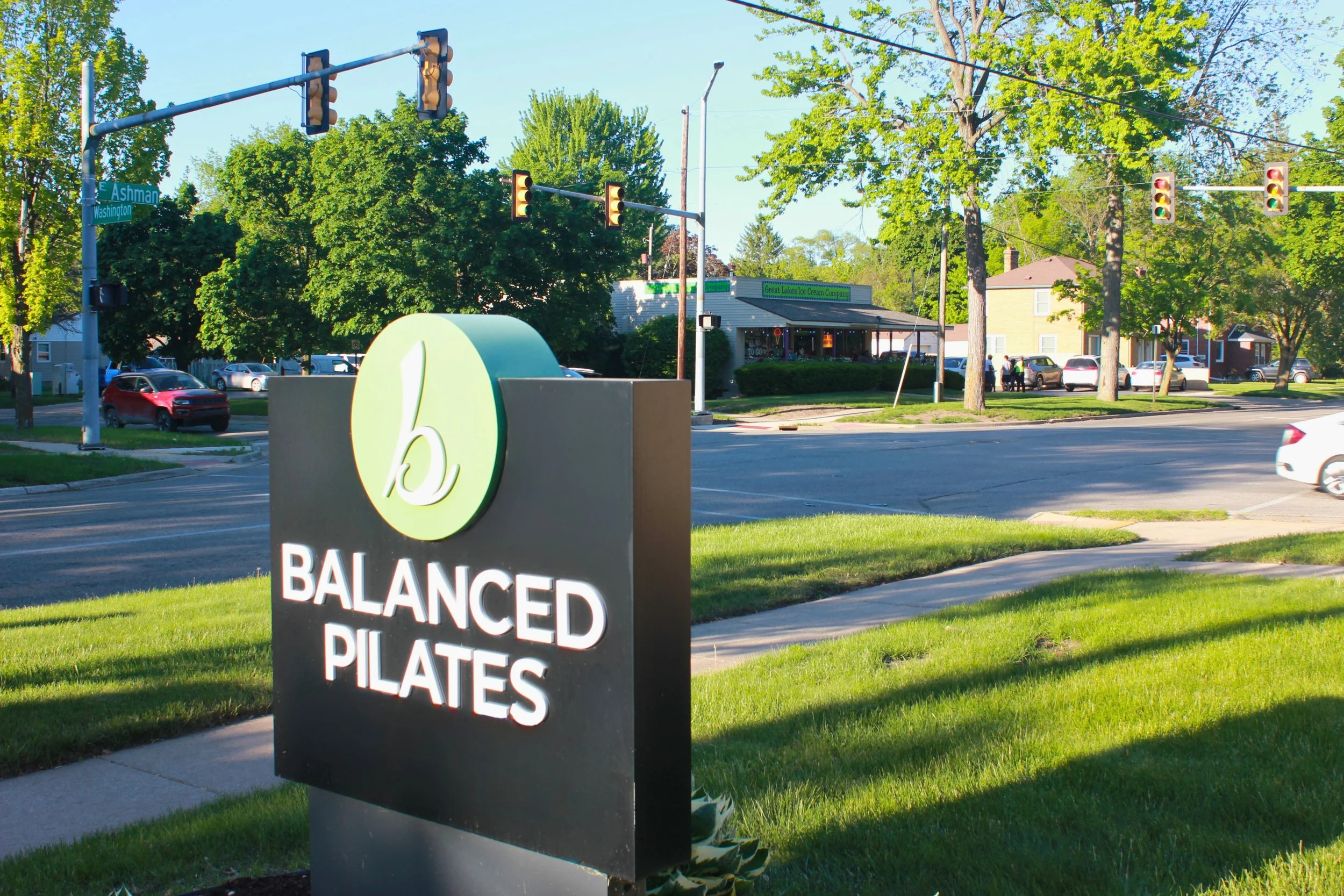 Street corner with traffic light, trees, a sign with the words 'Balanced Pilates', and a building with a green sign that reads 'Great Lakes Ice Cream Company.' Cars are parked along the street and people are walking.
