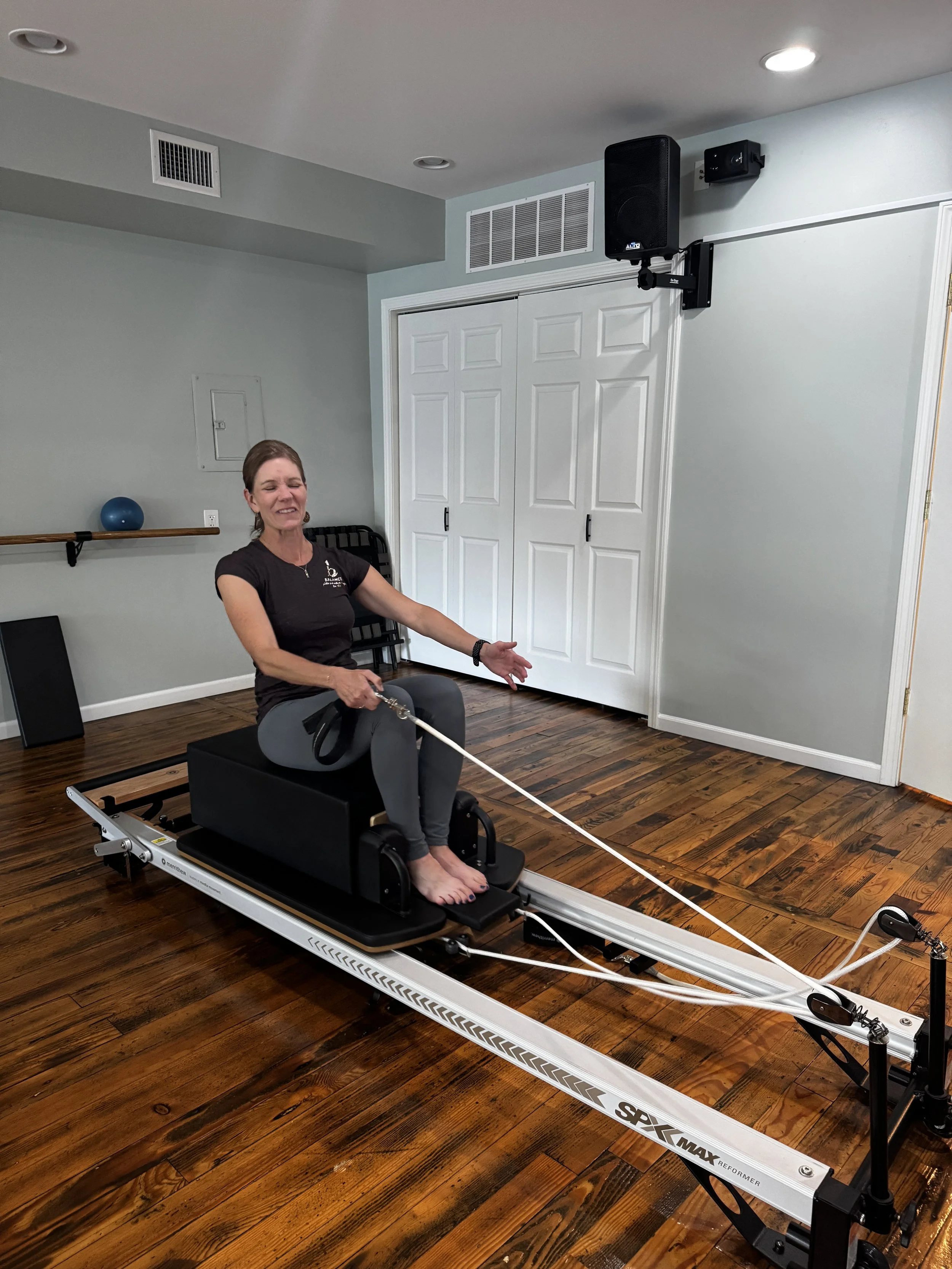 A woman on a Pilates reformer machine performing an exercise, in a room with a wooden floor, light-colored walls, and a white closet door at Balanced Pilates and Wellness Boutique in Midland, Michigan.