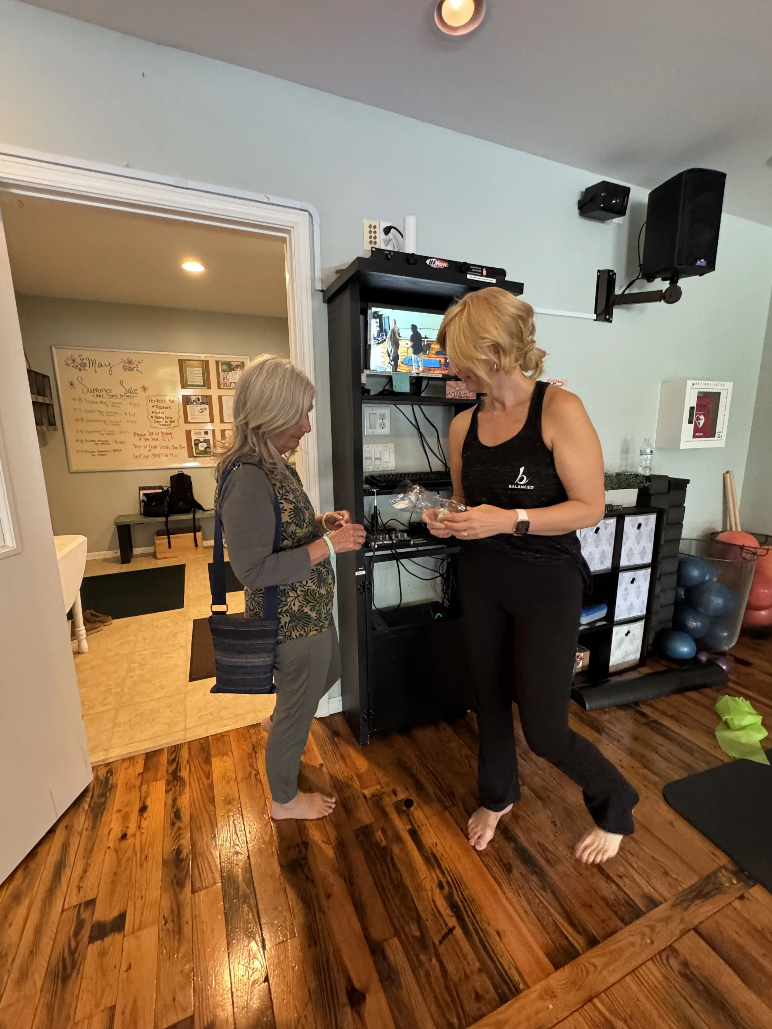 Two women barefoot inside a room, one with gray hair and wearing glasses, the other with blonde hair, appear to be engaging in a conversation or exchanging items near an electronic equipment shelf. There is a flat-screen TV on the shelf showing the s