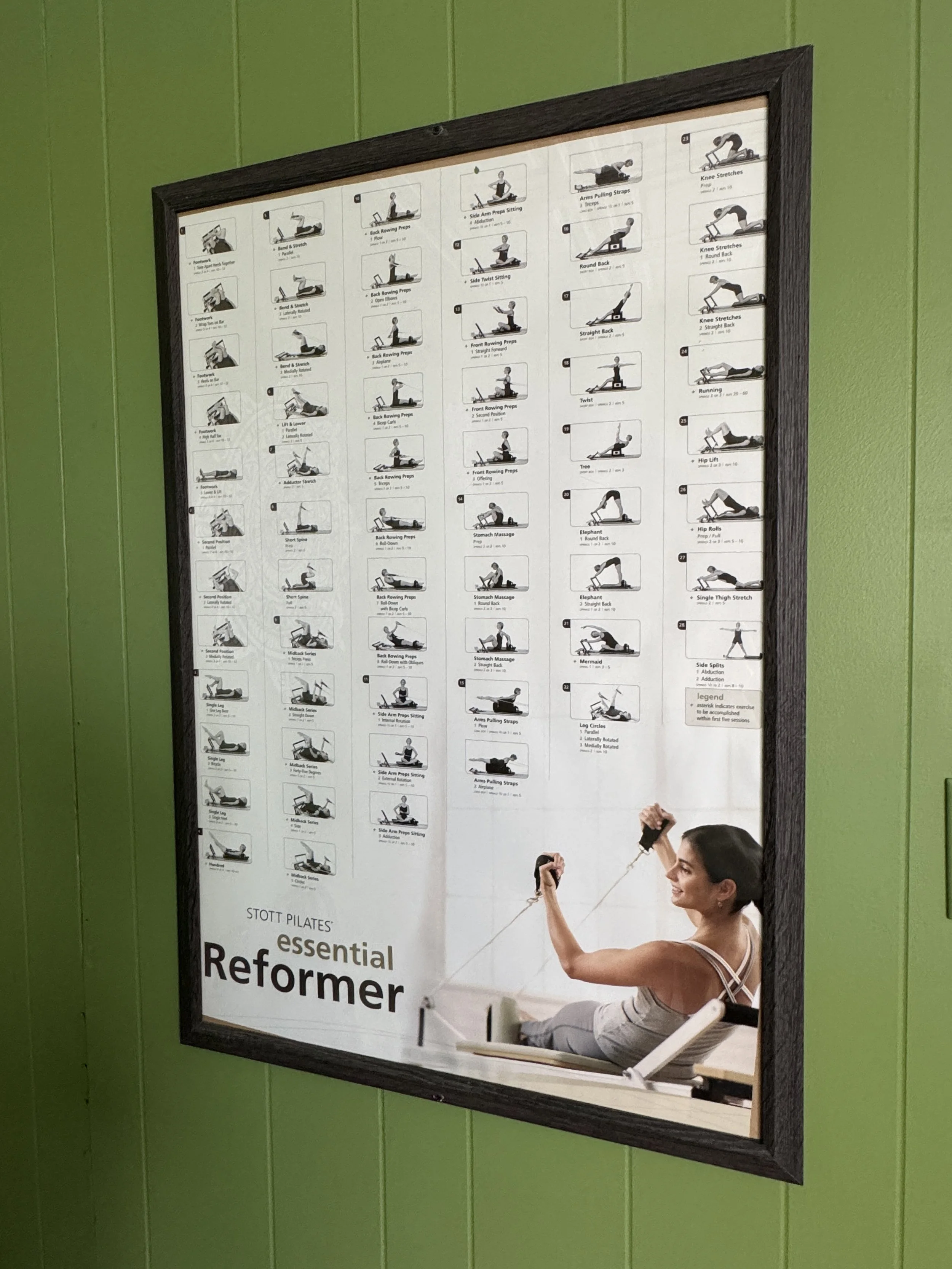 Framed poster showing various Pilates exercises with small illustrations and descriptions, on a green wall. A smiling woman with dark hair tied back is seated, holding resistance bands.