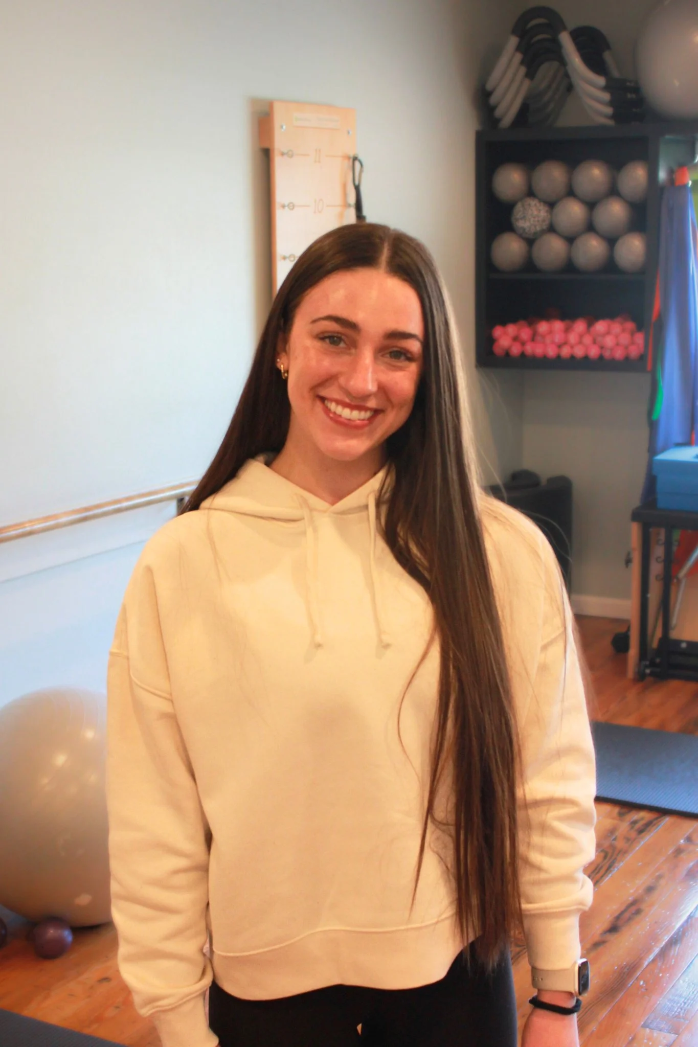 Pilates instructor Madeline Wenzell woman with long brown hair, smiling, wearing a beige hoodie and black pants, standing in a Pilates studio including medicine balls, kettlebells, and exercise mats in the background.