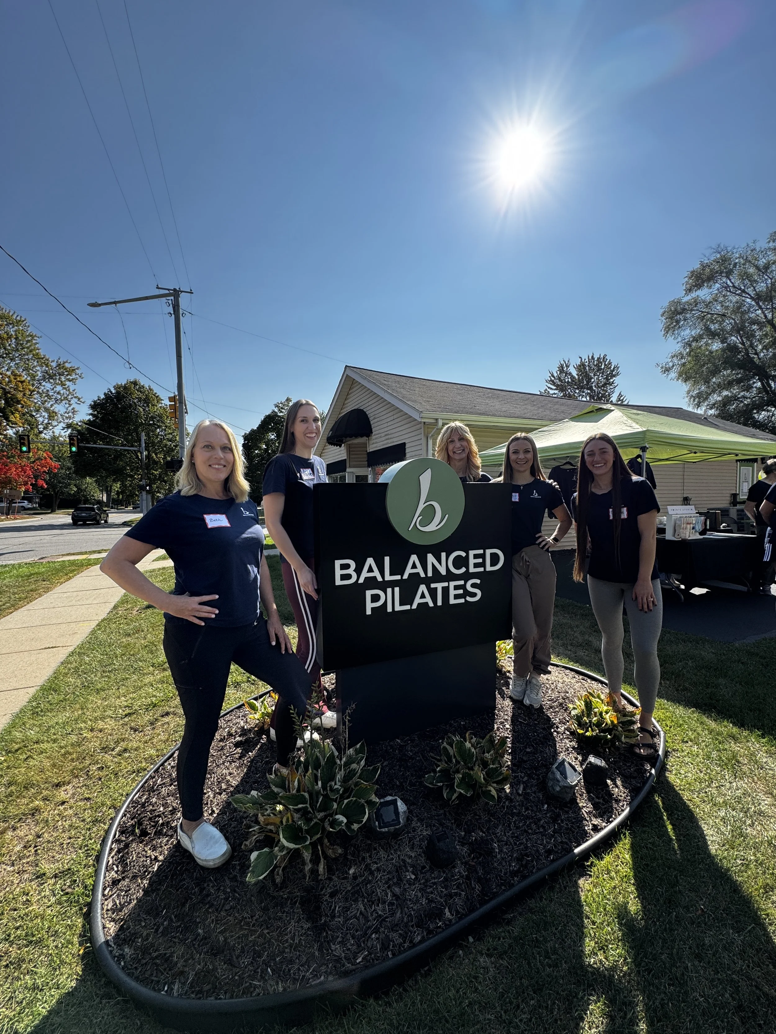 The Balanced team smiling outdoors at a community event, standing behind a sign that reads 'Balanced Pilates,' with a garden bed filled with plants and flowers, and a tent nearby on a bright sunny day.