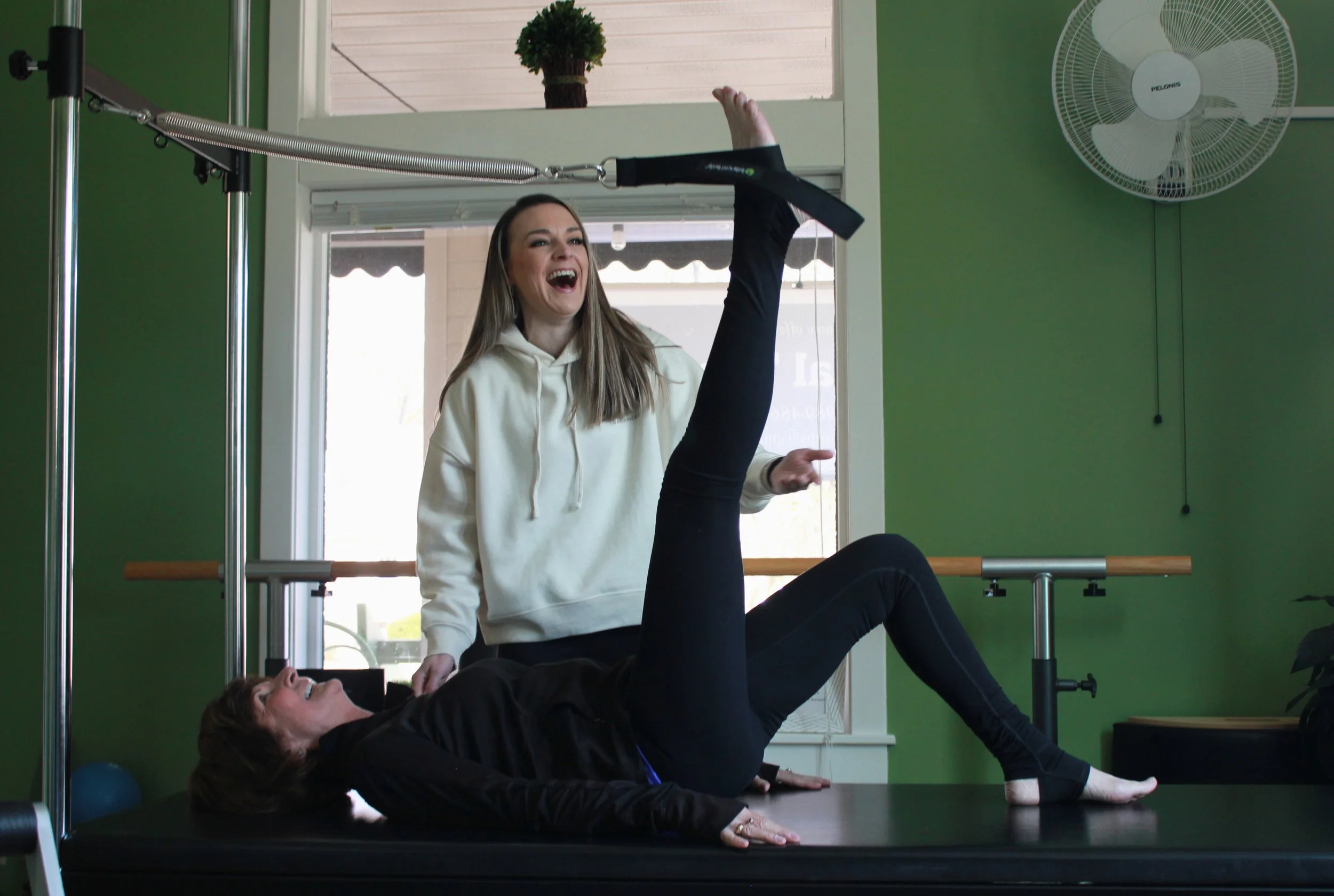 A woman is assisting a person with a pilates exercise using a reformer machine inside a green-walled room with a window, a fan, and a potted plant in the background.