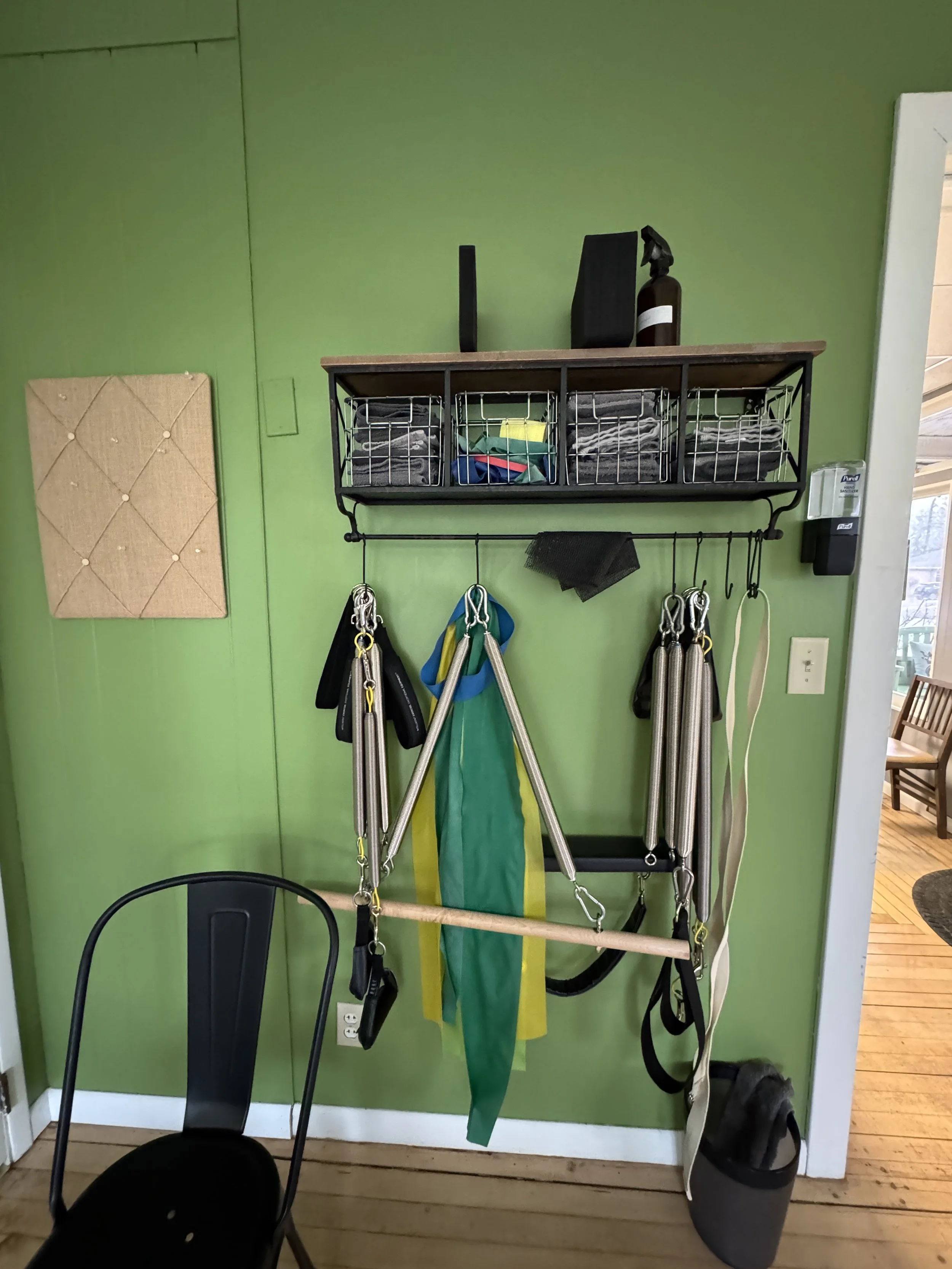 Pilates equipment storage with a black metal wall-mounted shelf holding spray bottles and folded towels, hooks with harnesses and leashes hanging, and rubber mats on a green wall with a beige cushion panel and a black chair in front.