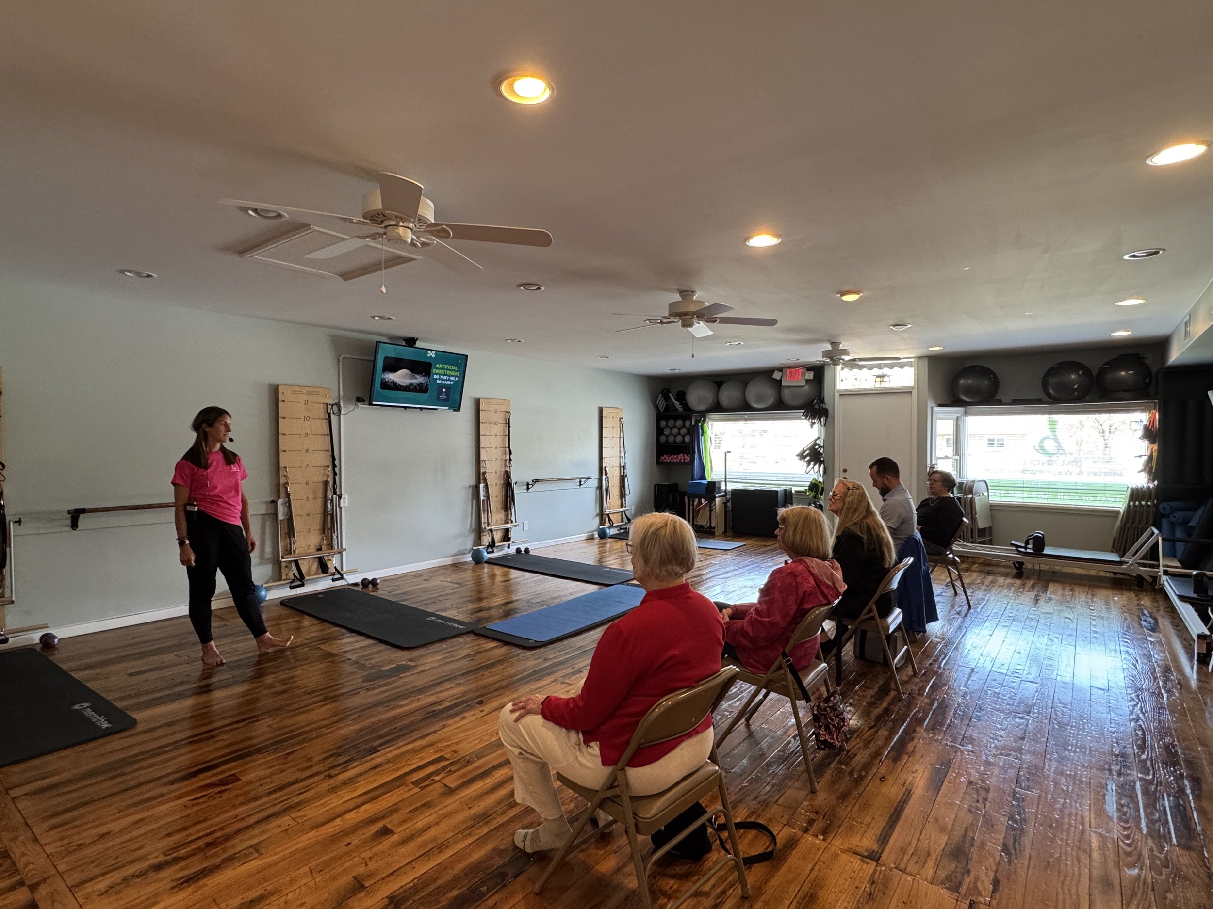People seated in chairs in a fitness studio for an educational workshop while an instructor, wearing a pink shirt, stands nearby. The room has hardwood floors and walls with fitness equipment and a television screen.